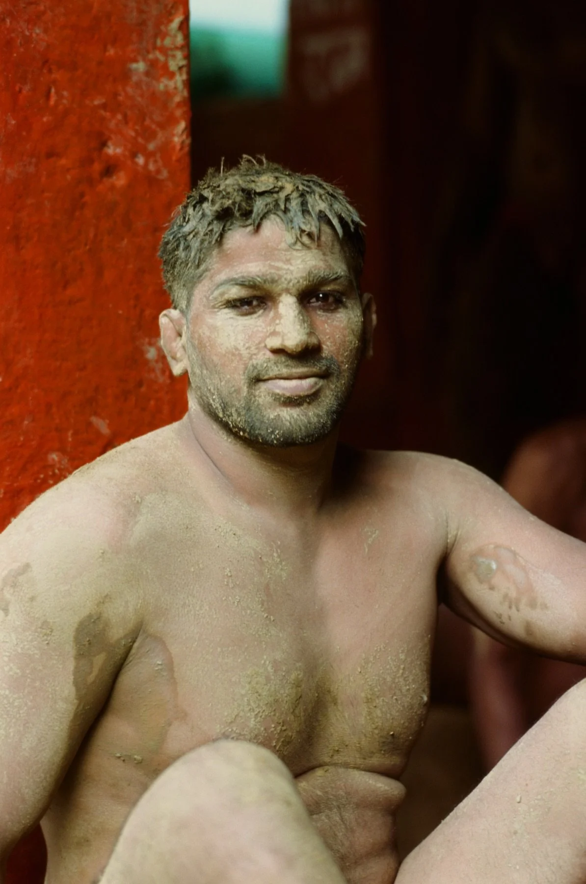 A shirtless man with dirty, dusty skin, sitting against a red wall, smiling at the camera.