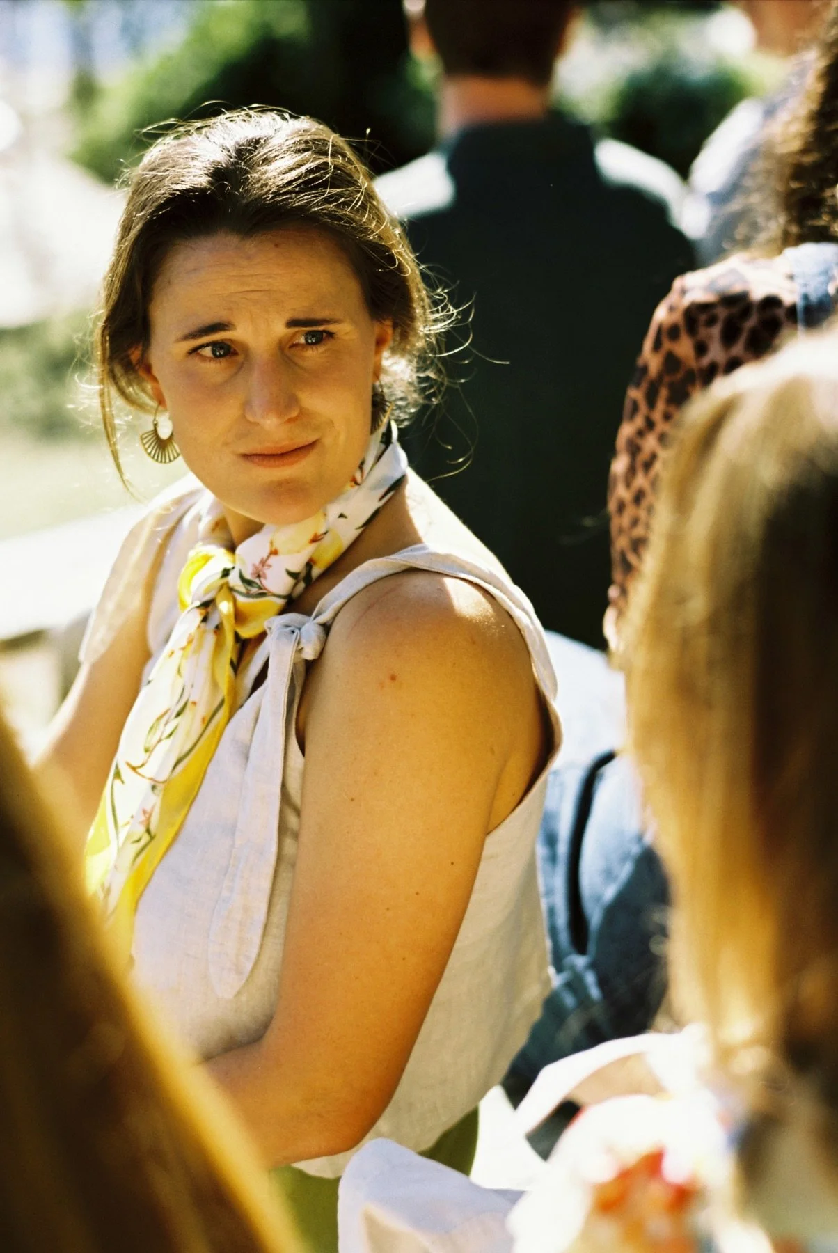 A woman with short brown hair, wearing a sleeveless white top and a yellow floral scarf, looking over her shoulder and smiling. She is outdoors in a sunny environment, with blurred figures behind her.