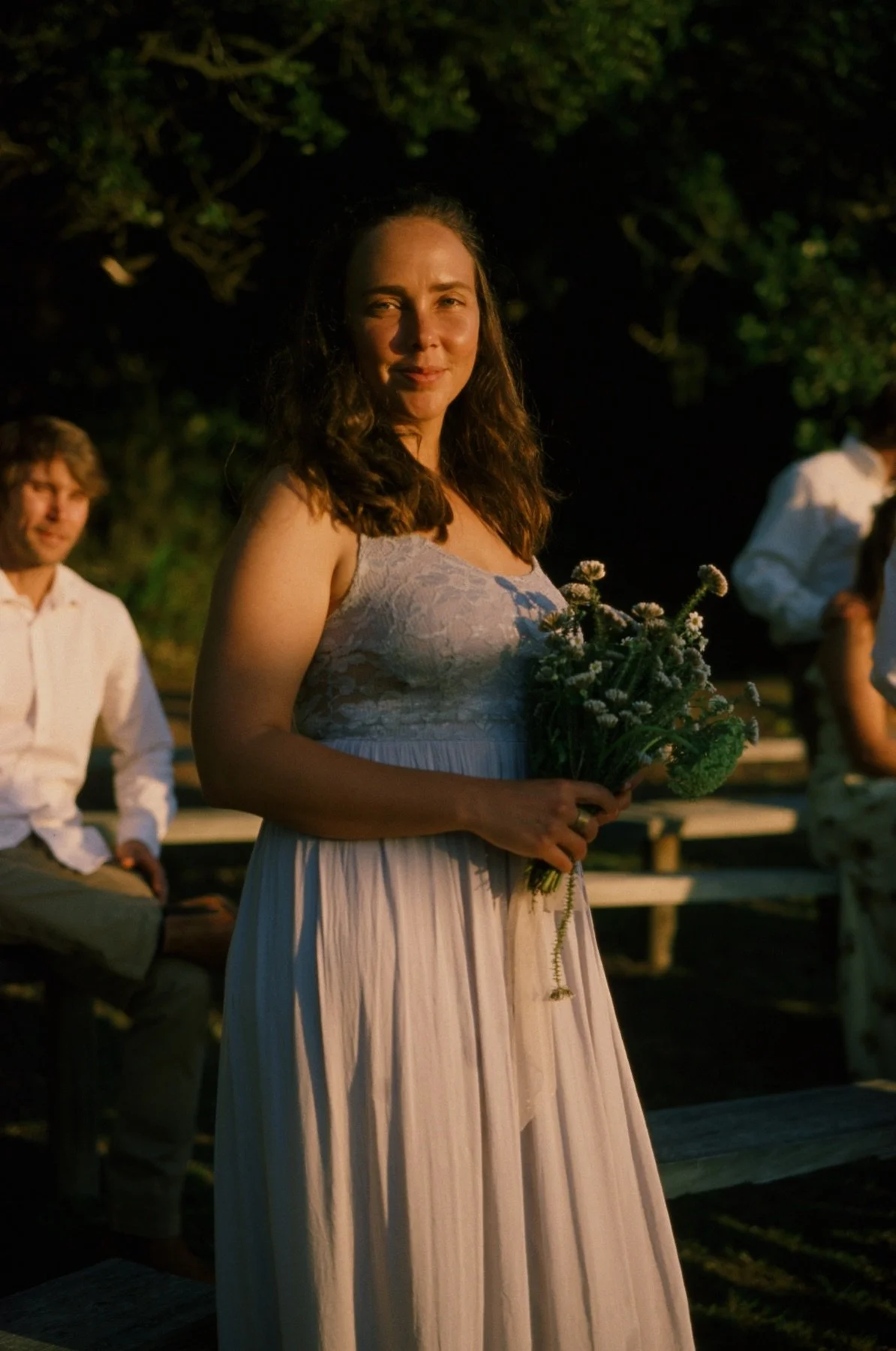A woman in a white dress holding a bouquet of flowers, standing outdoors at night during a gathering or celebration with seated people in the background.
