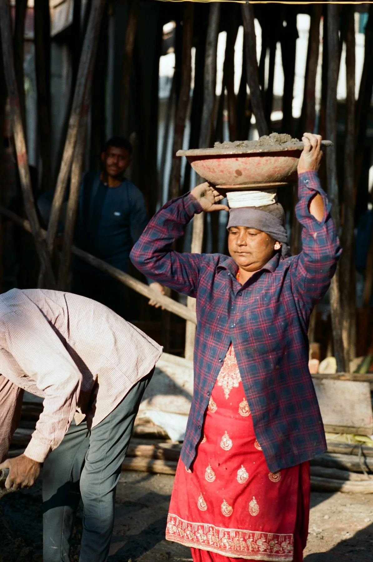 A woman wearing a plaid shirt and red traditional dress, balancing a basket of cement on her head, in a construction area. In the background, there is a man and wooden structures.