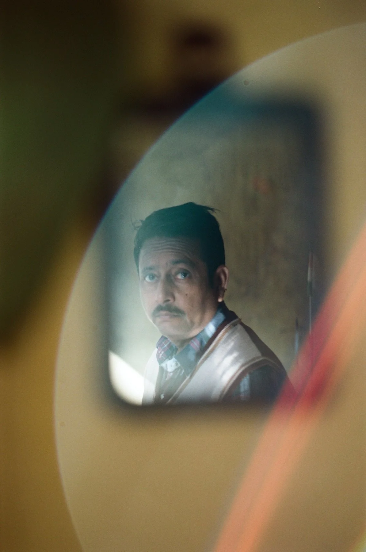 A man with dark hair and a mustache, wearing a plaid shirt and jacket, looking into a mirror or reflective surface.