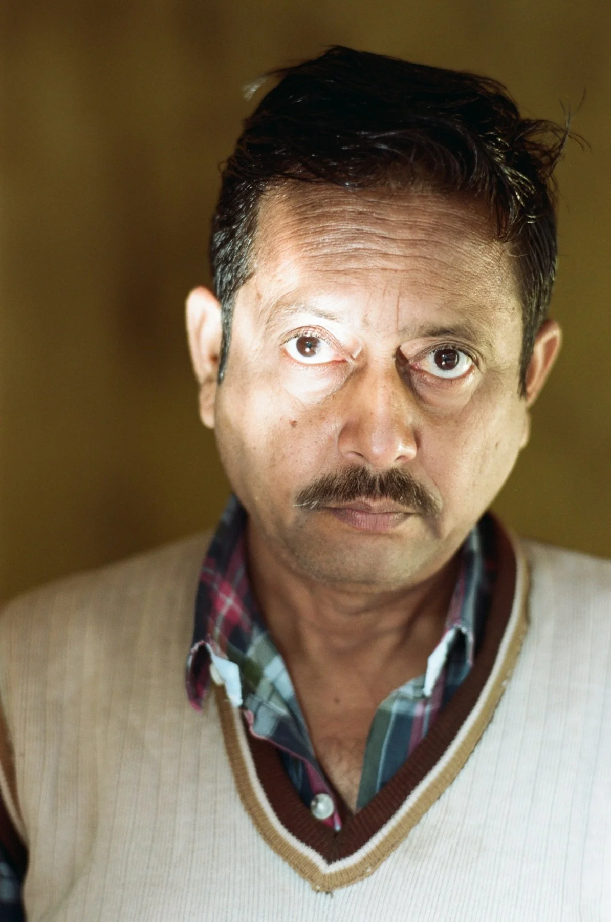A man with short dark hair, a mustache, and light skin wearing a beige sweater over a plaid shirt, looking directly at the camera with a serious expression.