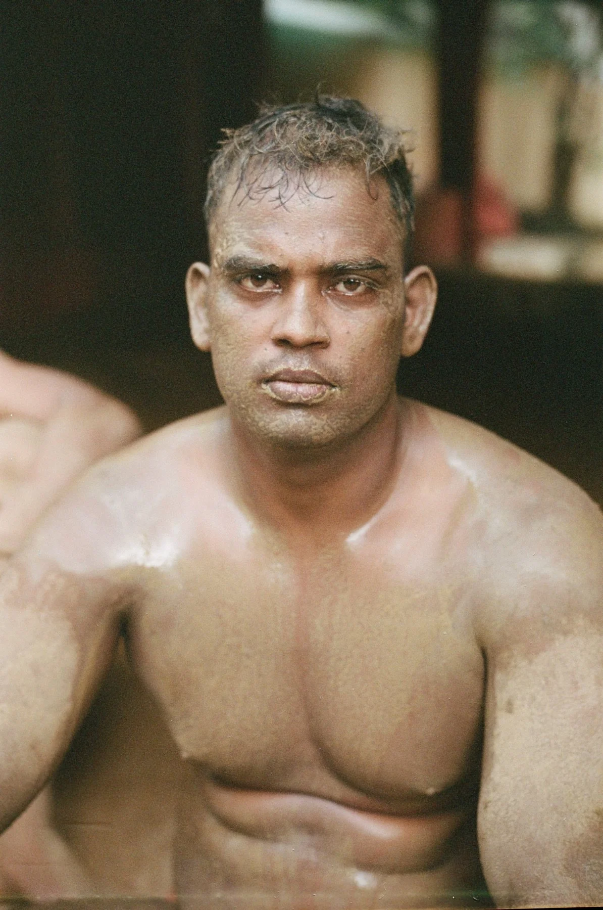A shirtless man with short, wet hair sits in a bath or hot spring, looking directly at the camera with a serious expression.
