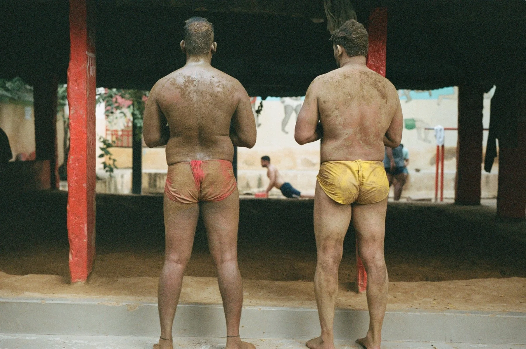 Two male wrestlers with dirt on their backs, standing in a wrestling ring with red posts, viewed from behind, in a gymnasium with children playing in the background.