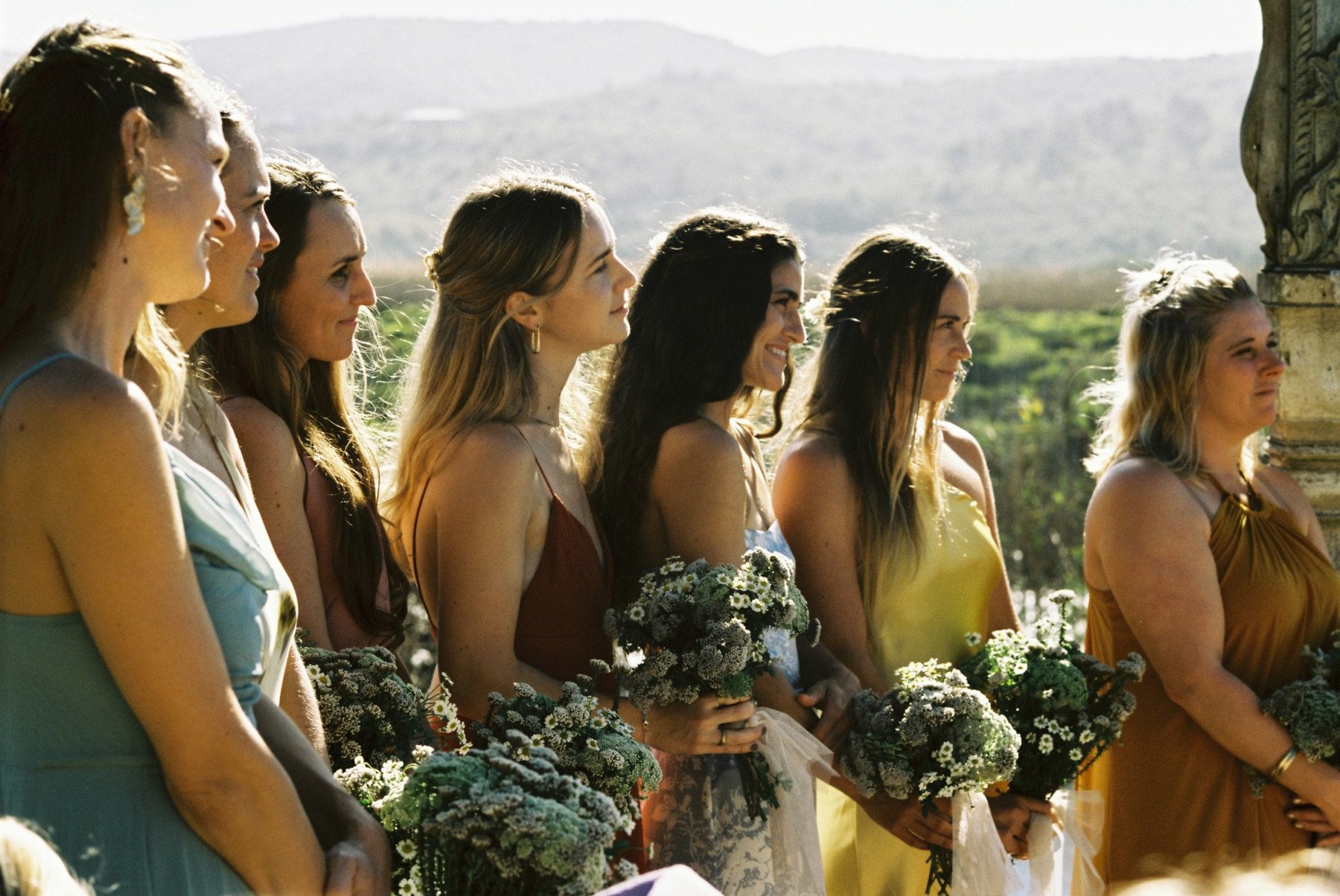 A group of women with flowers, standing in a line outdoors during daylight, possibly at a wedding or celebration.
