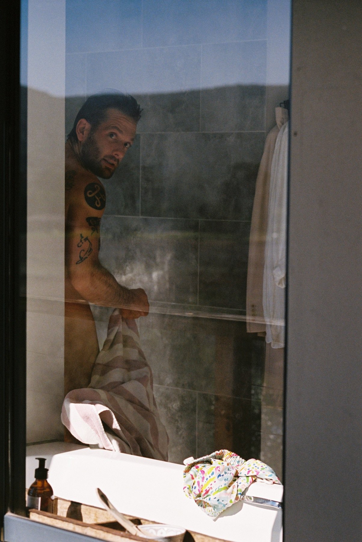 A man with tattoos on his upper arm and shoulder, looking into a shower with a glass door, holding a striped towel. The shower has gray tile walls, with a towel hanging on a hook. In the foreground, there is a white sink with a bottle of soap and a c