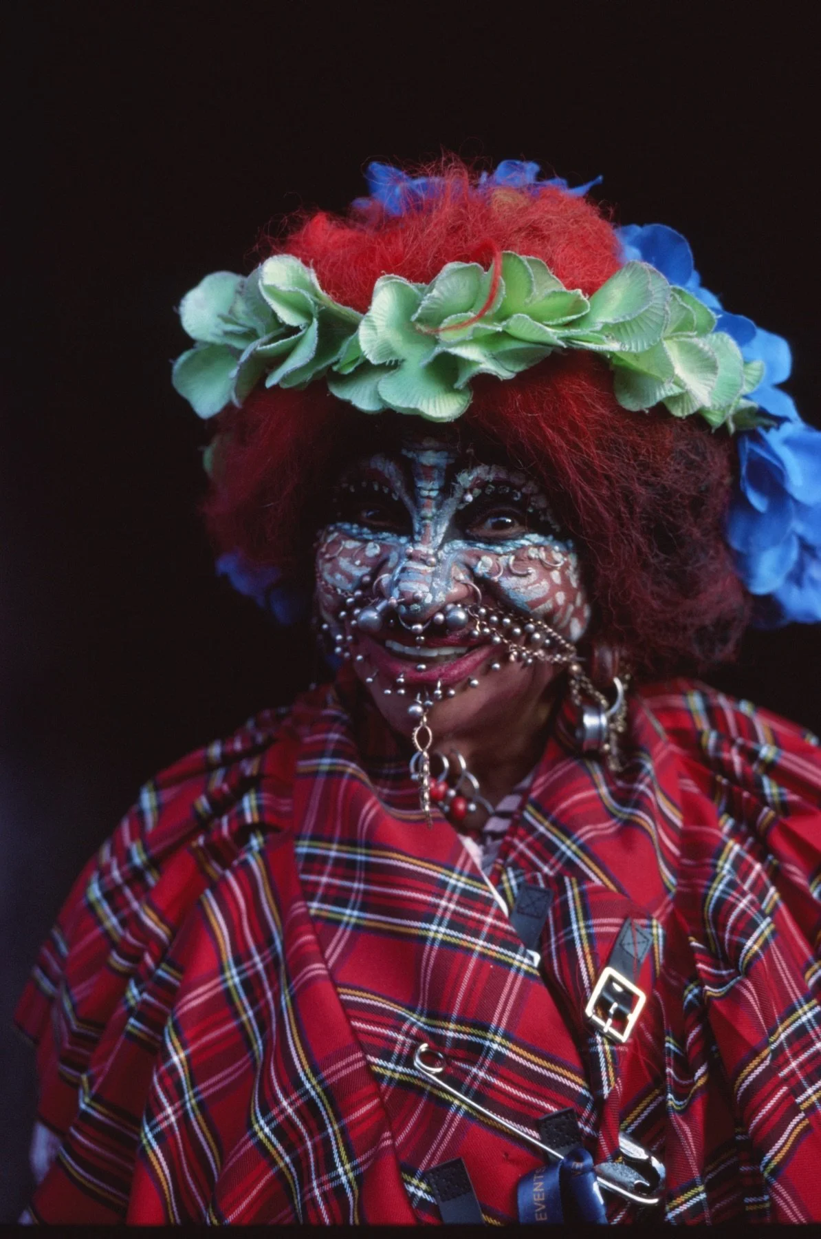 A person smiling, wearing face paint, jewelry, a red plaid shirt, and a colorful flower crown with blue and green flowers. They have red hair and are set against a dark background.