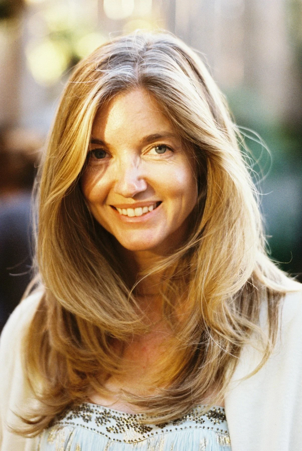 A smiling woman with long, wavy, blonde hair and blue eyes, wearing a white top with embellishments around the neckline.