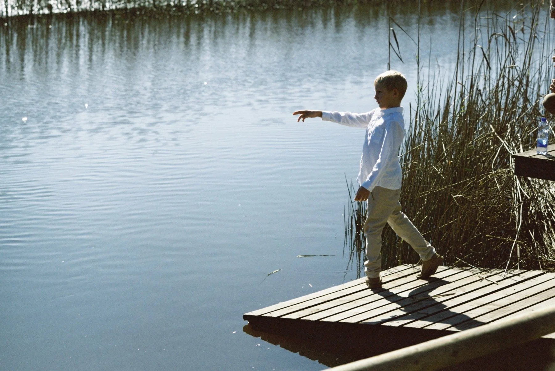 A young boy in white shirt and khaki pants standing on a wooden dock, reaching out towards the water, with reeds and a bottle on a small table nearby, by a calm lake.