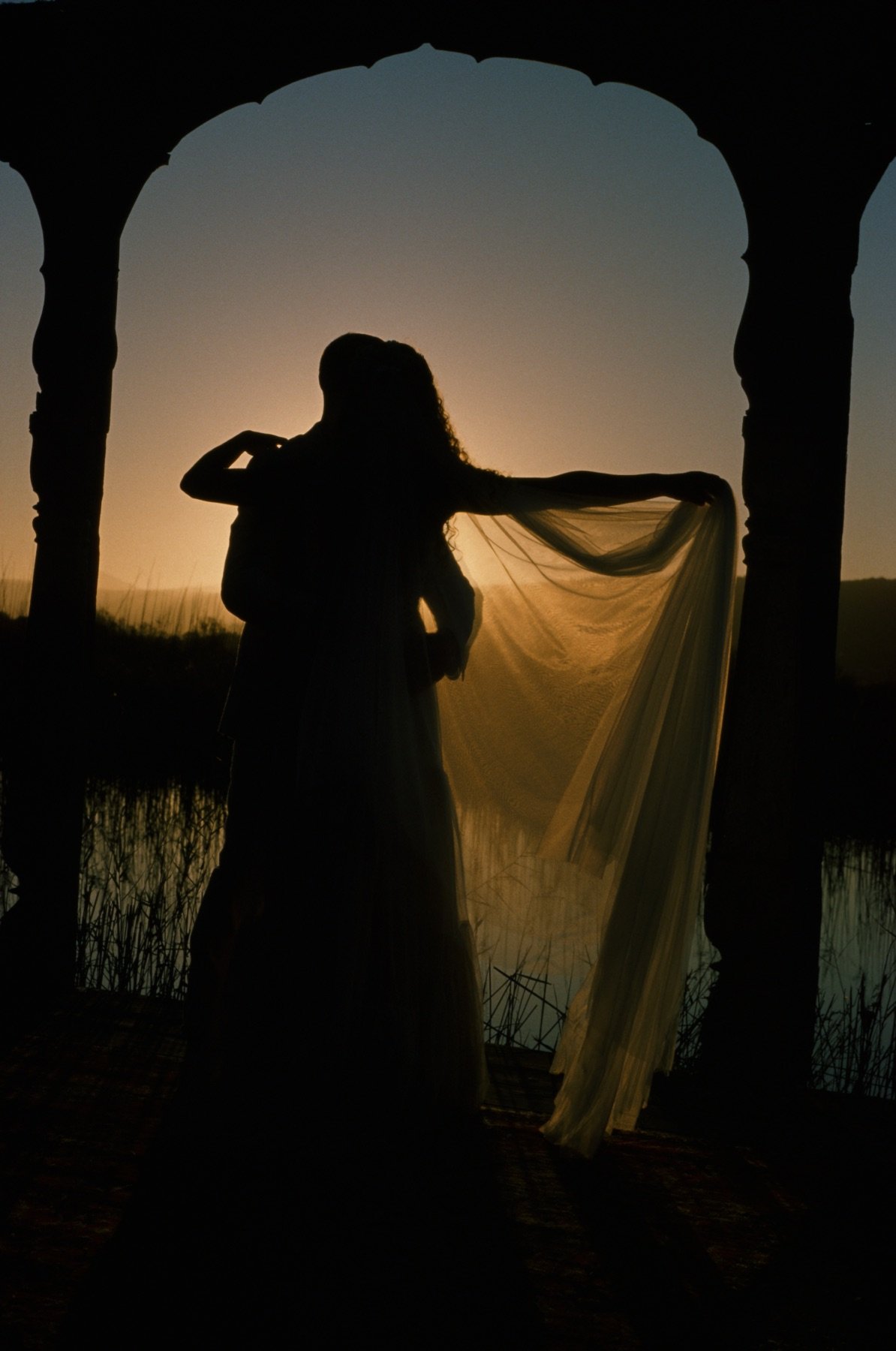Silhouettes of a couple embracing and a woman holding a translucent fabric, standing beneath an arched structure at sunset or sunrise over water.