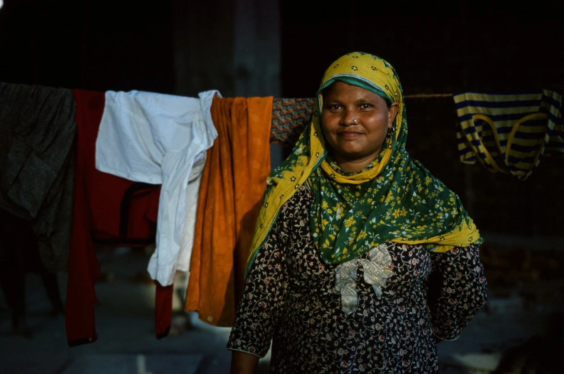 A smiling woman wearing a yellow and green headscarf stands indoors, with colorful laundry hanging behind her.