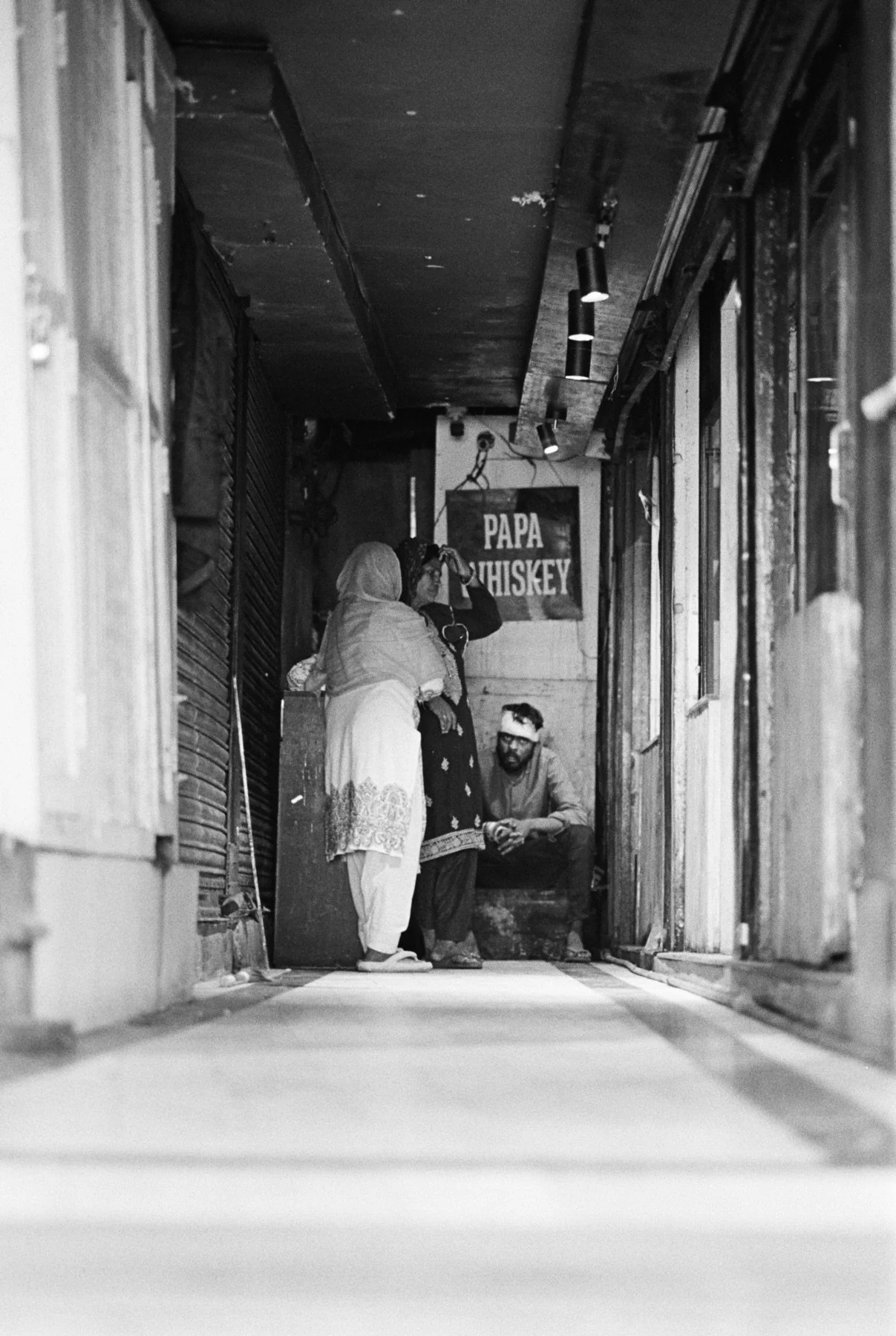 A black and white photo of a narrow alleyway with three people engaged in conversation. A man with a headband is sitting on a ledge, while two women, one wearing a hijab, stand nearby. A sign that reads 'PAPA WHISKEY' hangs on the wall in the backgro
