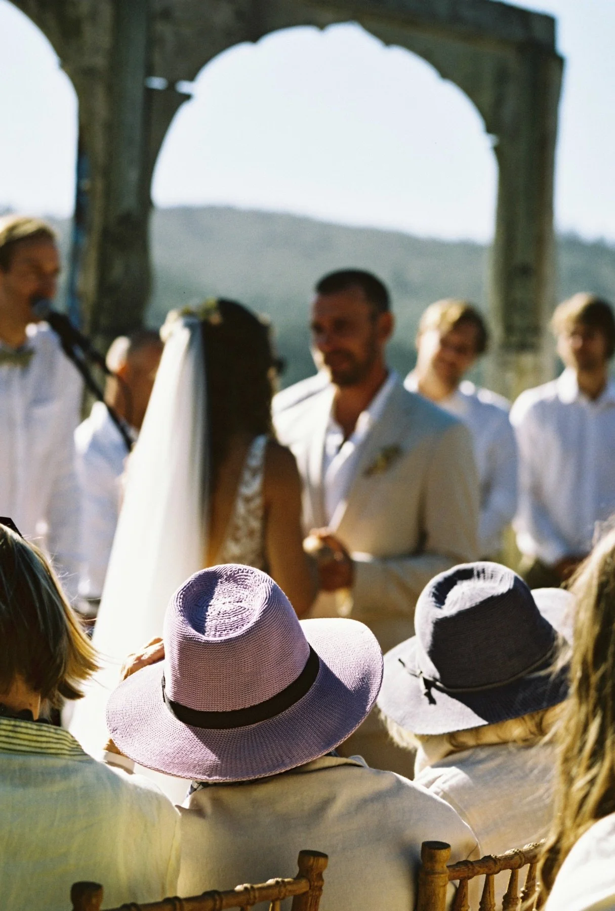 A wedding ceremony outdoors with a couple exchanging vows in front of an arch, surrounded by guests wearing hats.