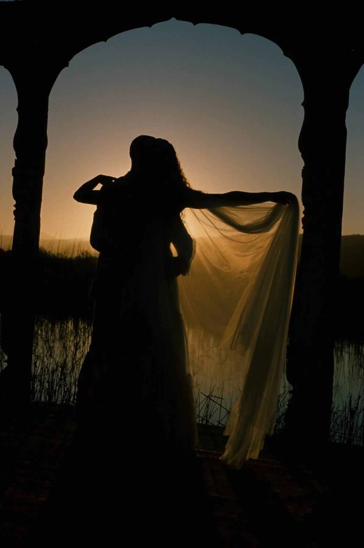 Silhouette of a woman holding a flowing fabric near a body of water at sunset, framed by an arched stone structure.