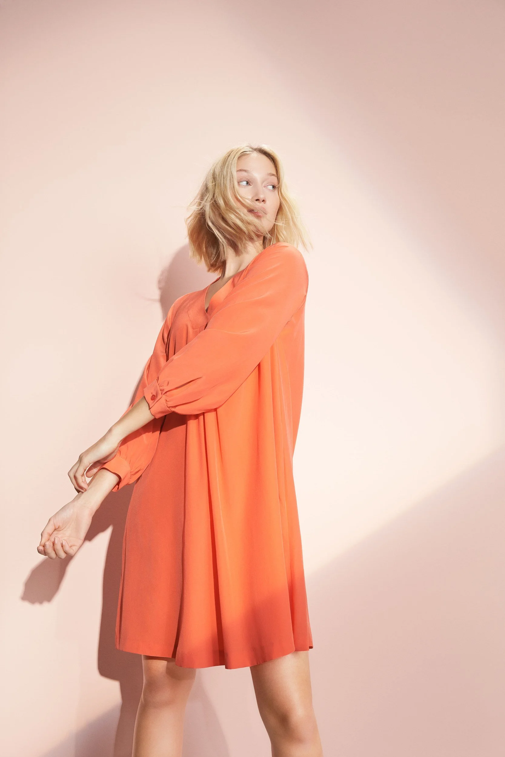 Hellmuth Lookbook SS20: Kleid DALLAS in Flamingo-Orange, stehend vor rosé Studiowand