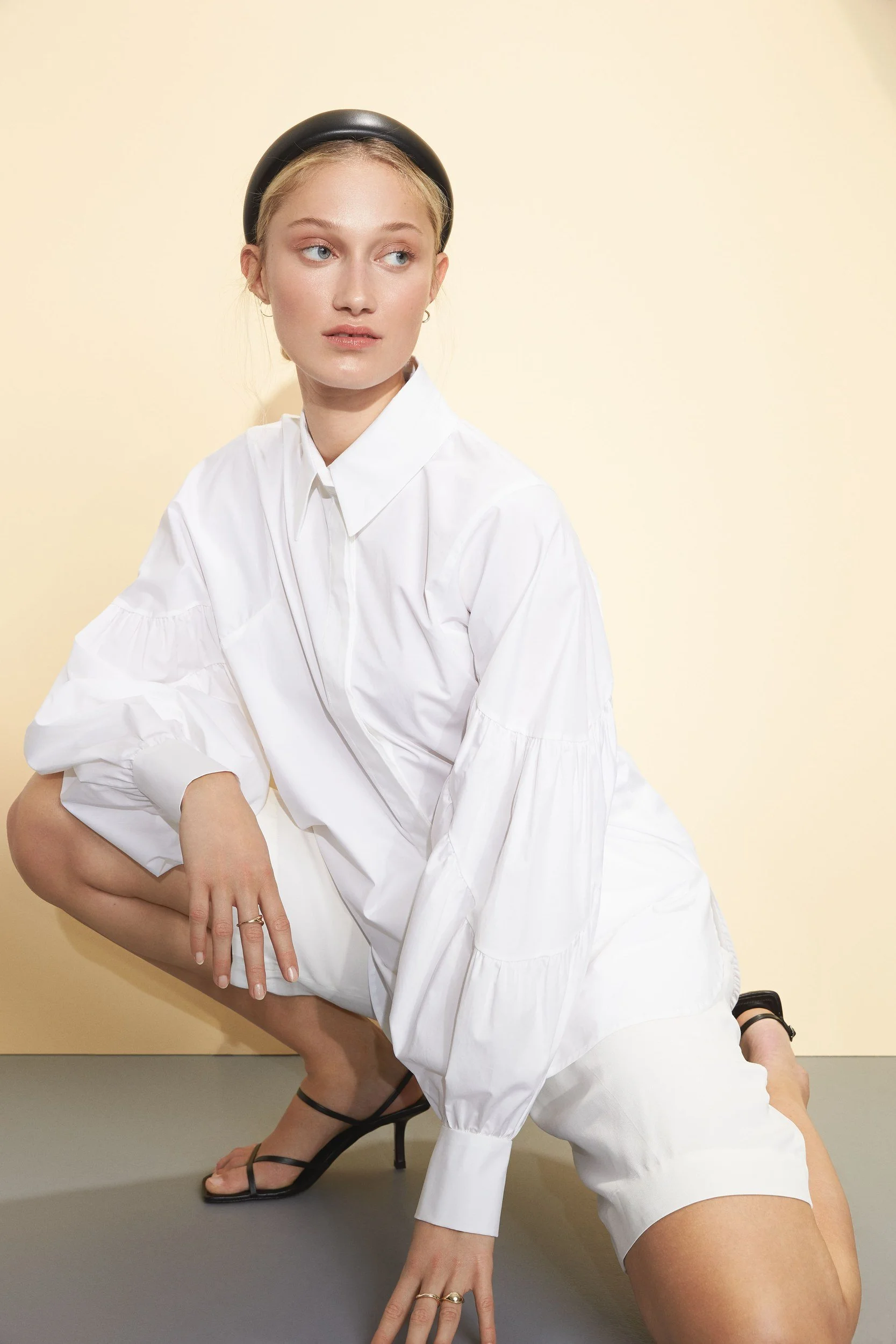 Hellmuth Lookbook SS20: weißes Blusenkleid BELLA, hockende Pose vor Vanille-Wand