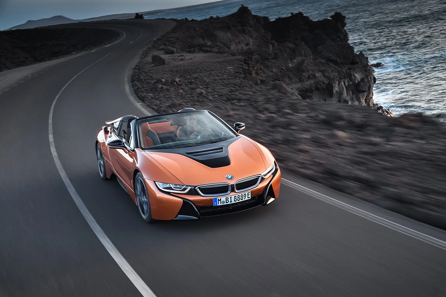 Oranger BMW i8 Roadster auf Küstenstraße vor Lavafelsen und Meer in der Dämmerung – Retusche Bildergut.