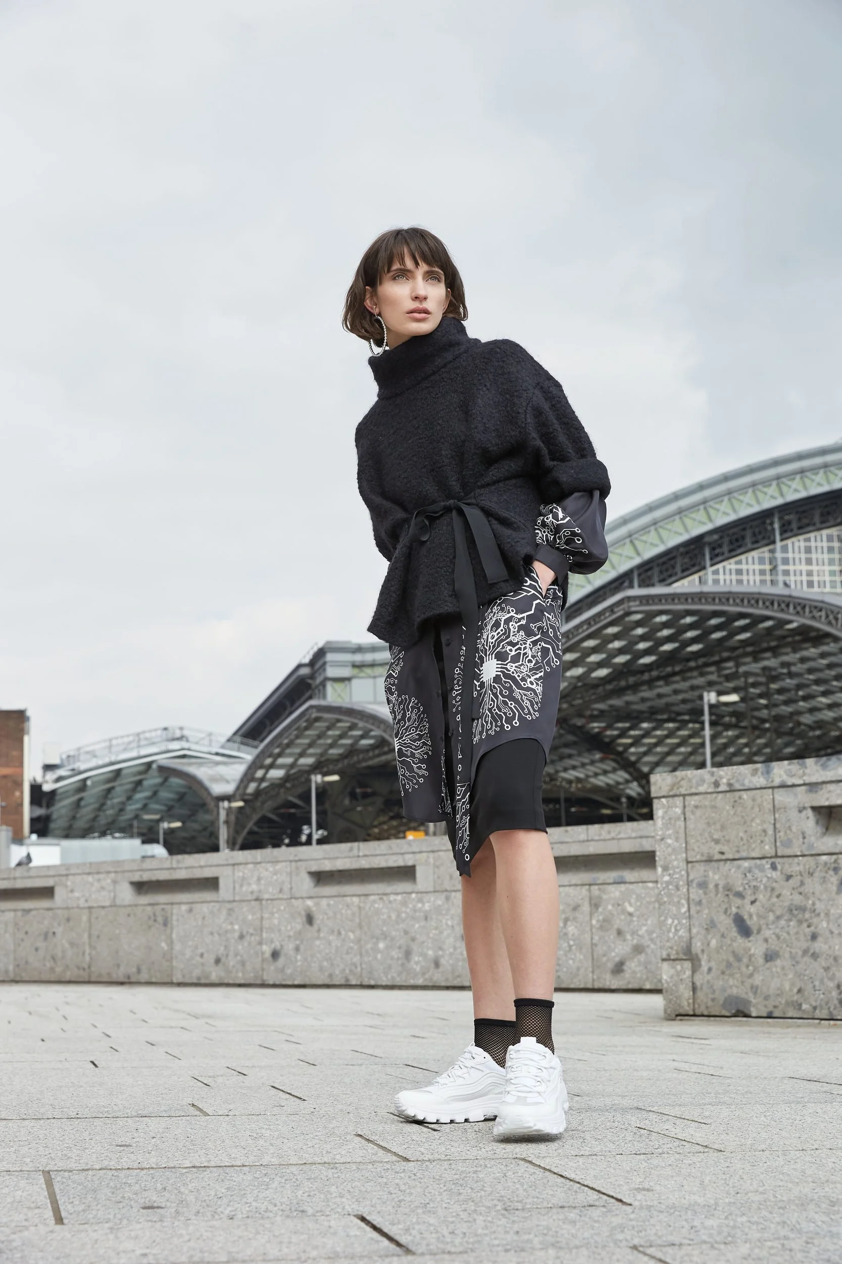 Hellmuth Lookbook 2019: schwarzer Look KUNIS mit gemustertem Rock vor dem Dach des Kölner Hauptbahnhofs
