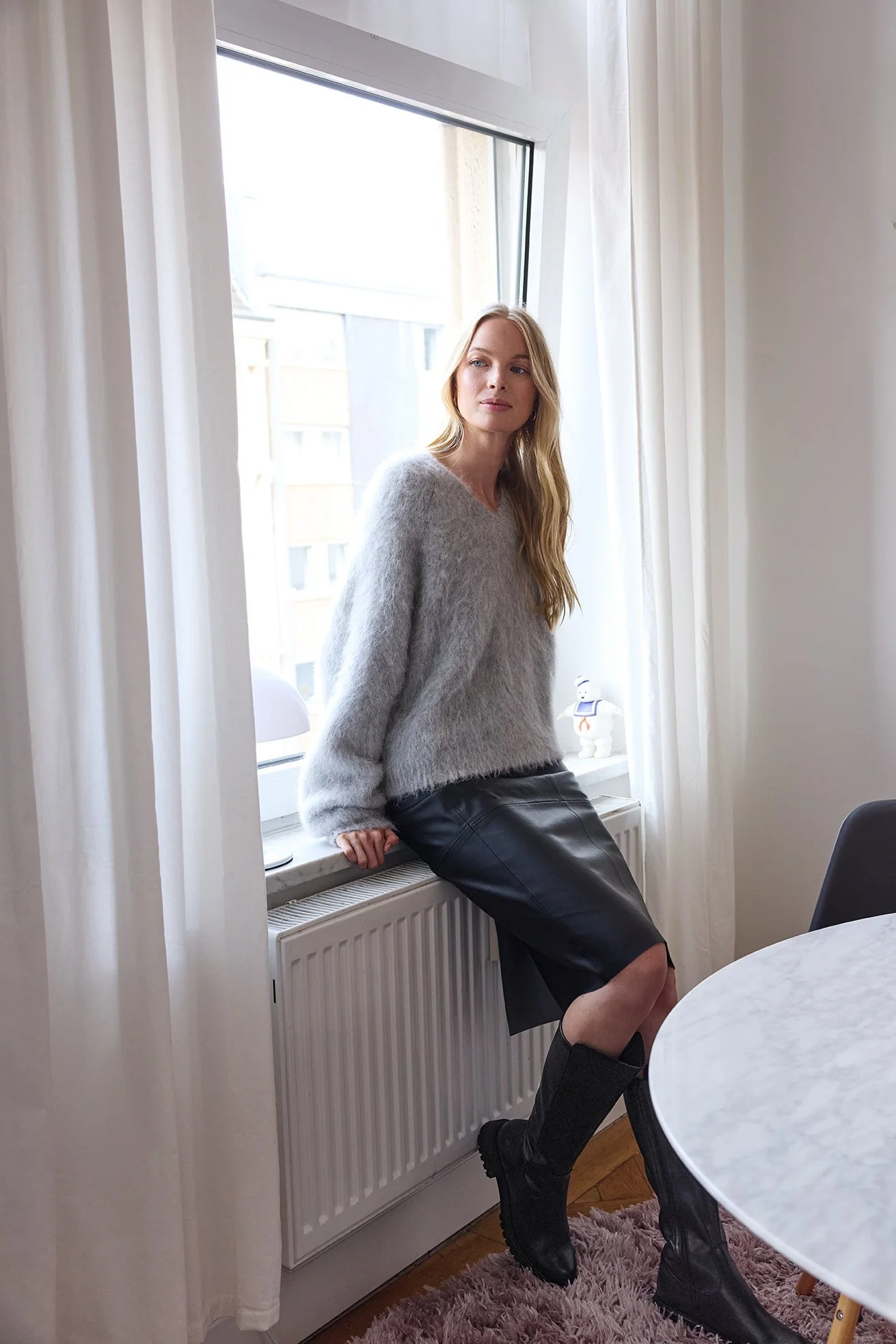 Blondes Model in grauem Mohairpulli und Lederrock am Fenster, Trendkampagne 2024 für Zinser – Retusche Bildergut.