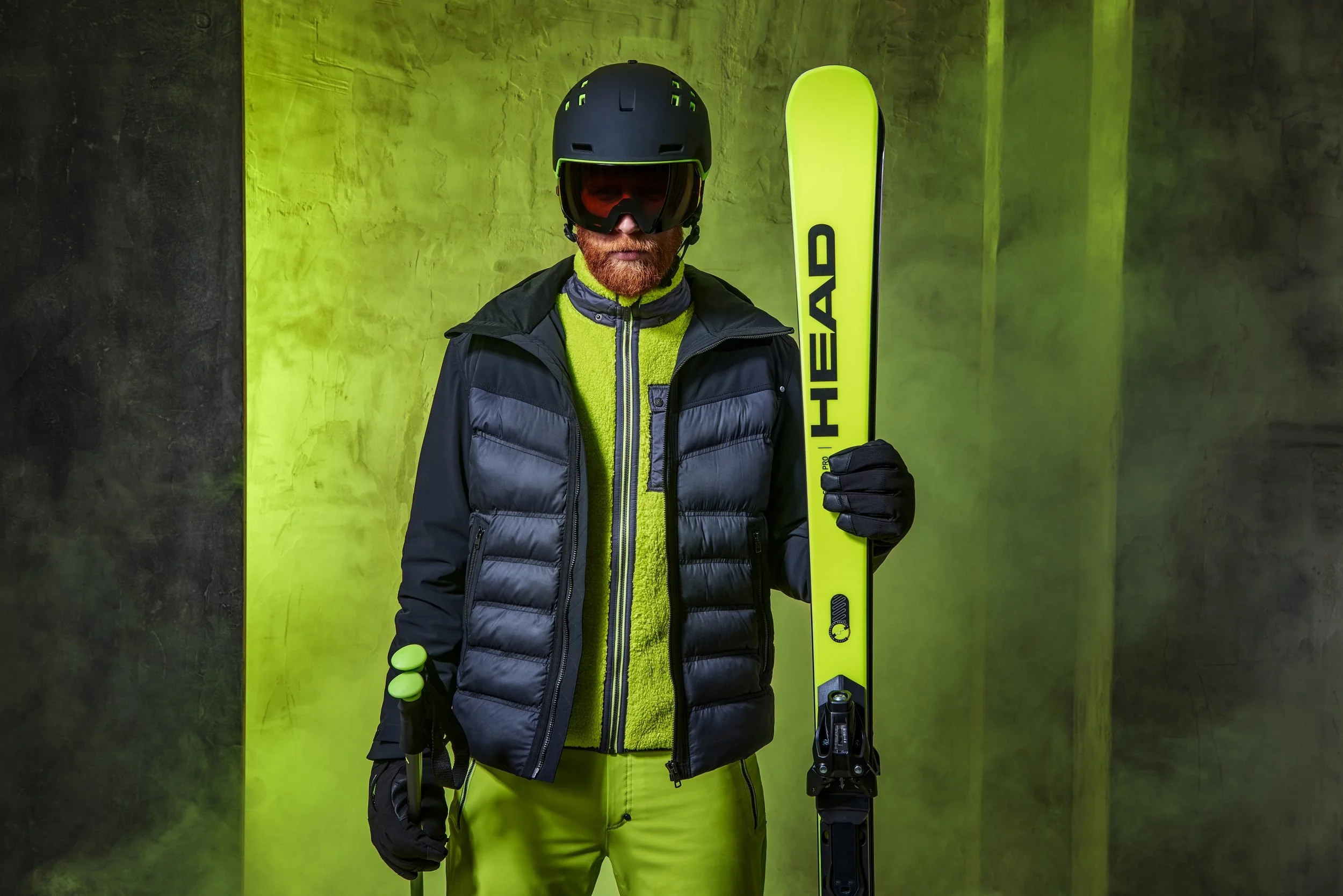 Mann in dunkler Daunenjacke vor giftgrüner Wand mit HEAD-Ski