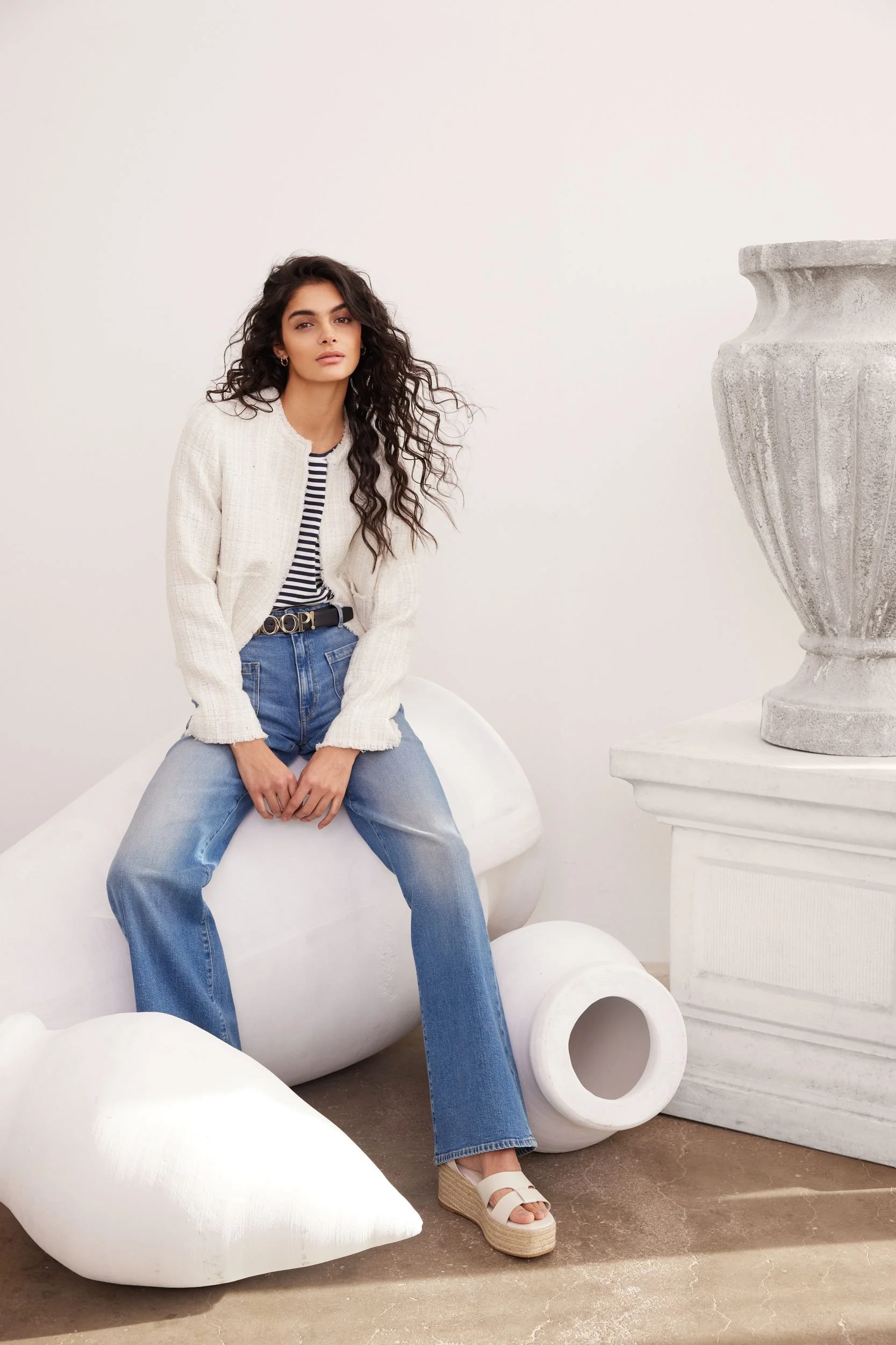 Modehaus Zinser Frühjahr-Sommer 2024: weiße Strickjacke über Streifenshirt und Jeans, sitzend auf liegender Vase