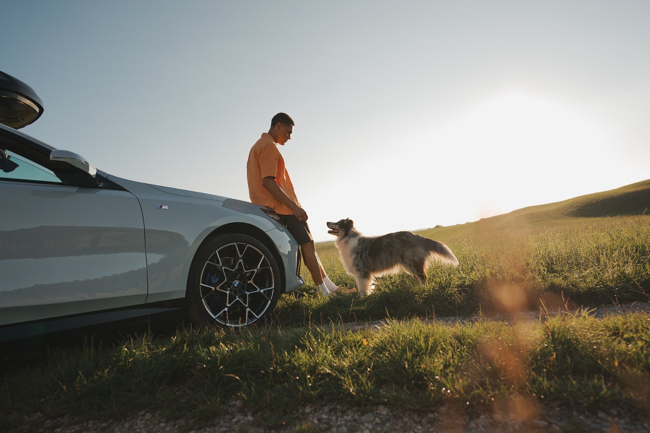 Mann lehnt an weißem BMW und begrüßt einen Hund im Abendlicht auf sommerlicher Wiese – Retusche Bildergut.