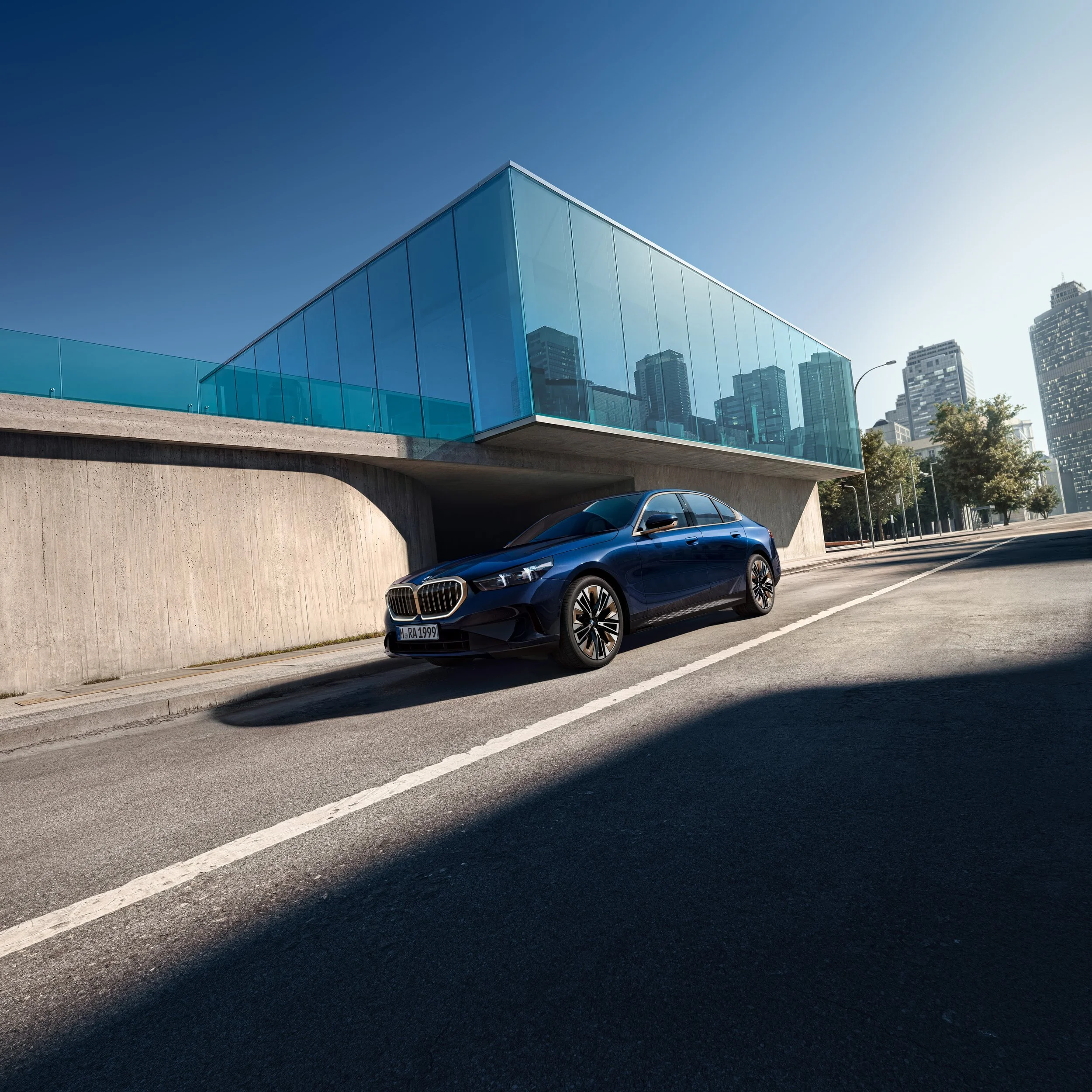 Dunkelblaue BMW 5er Limousine vor gläsernem Architekturbau mit Skyline bei Sonne – Retusche Bildergut.