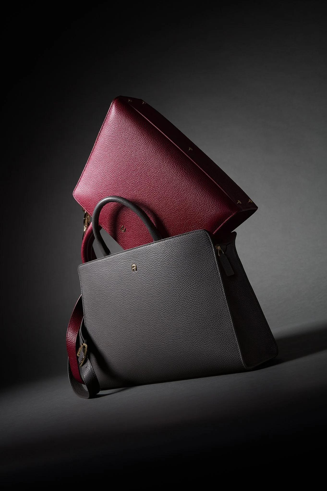 Bordeauxrote und graue Aigner-Ledertaschen im dramatischen Licht vor dunklem Hintergrund – Retusche Bildergut.
