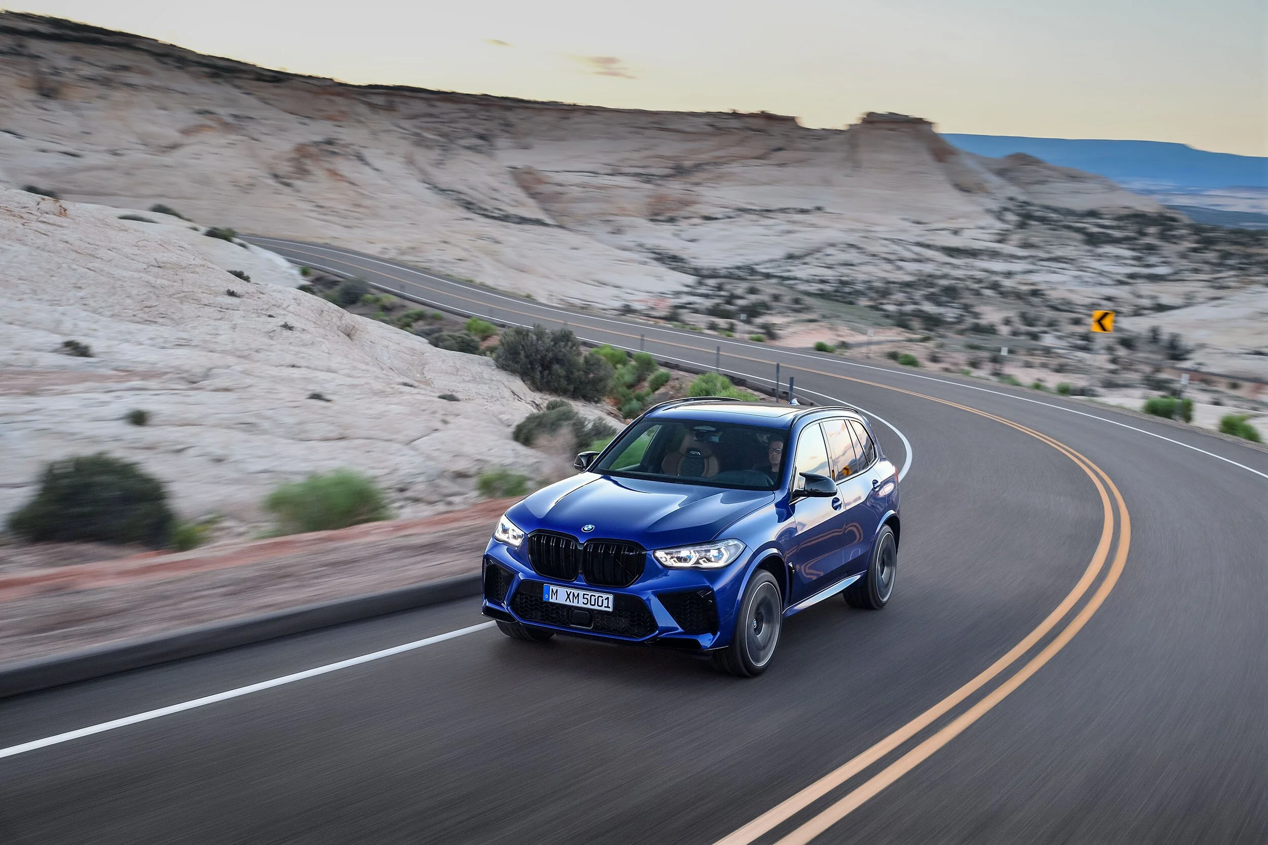 Blauer BMW X5M auf kurviger Straße vor heller Felslandschaft in der Dämmerung – High-End-Retusche von Bildergut.