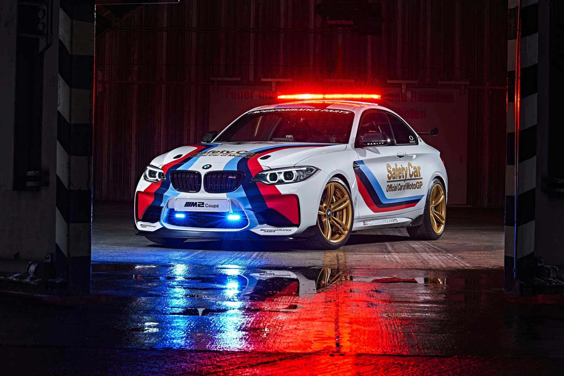 BMW M2 MotoGP Safety Car mit goldenen Felgen und Blaulicht in dunkler Halle – Retusche Bildergut.