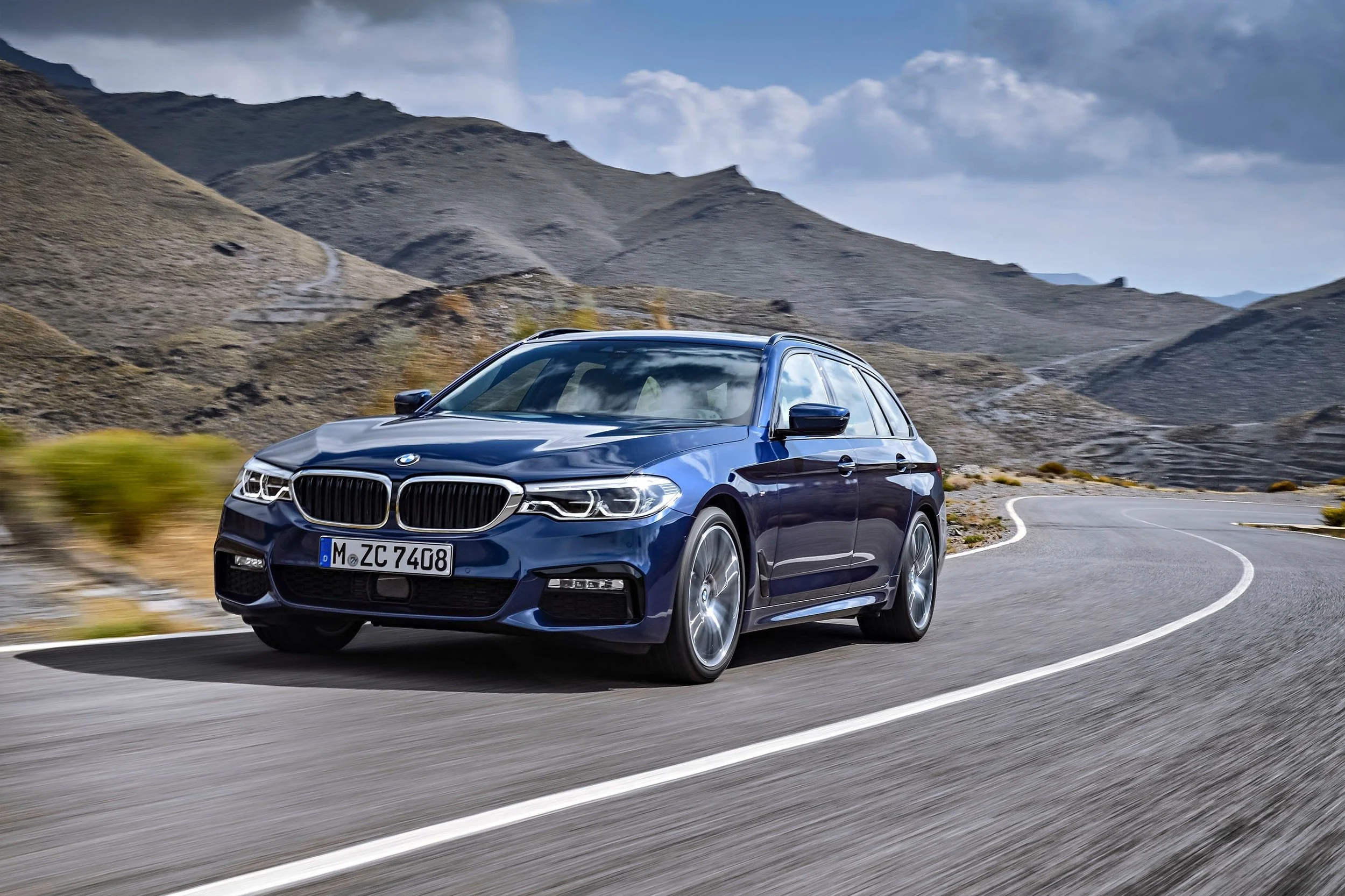 Blauer BMW 5er Touring in Fahrt auf kurviger Bergstraße vor karger Berglandschaft – Retusche Bildergut.