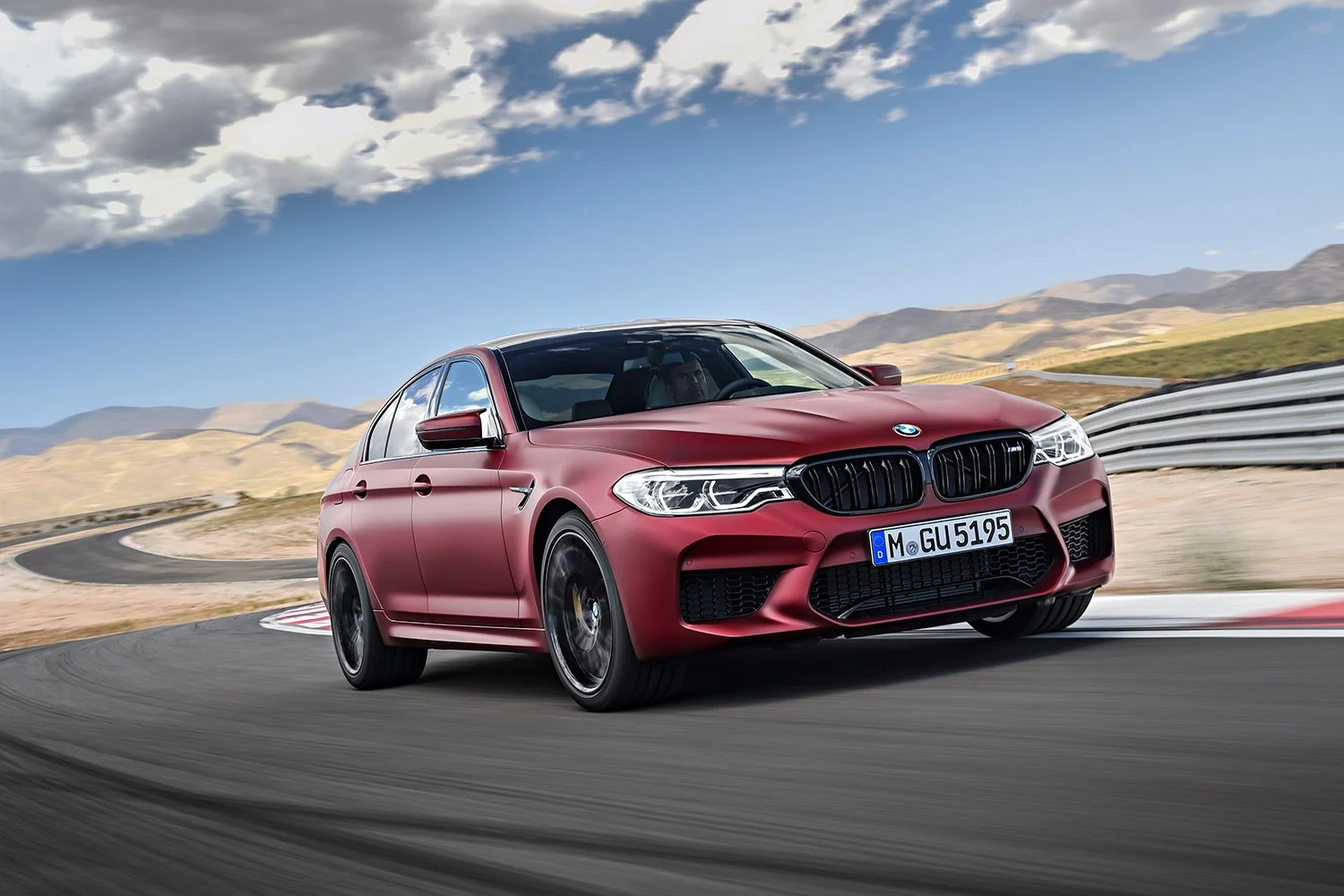 Mattroter BMW M5 Competition auf offener Fläche vor Bergen und Wolkenhimmel – Retusche Bildergut.