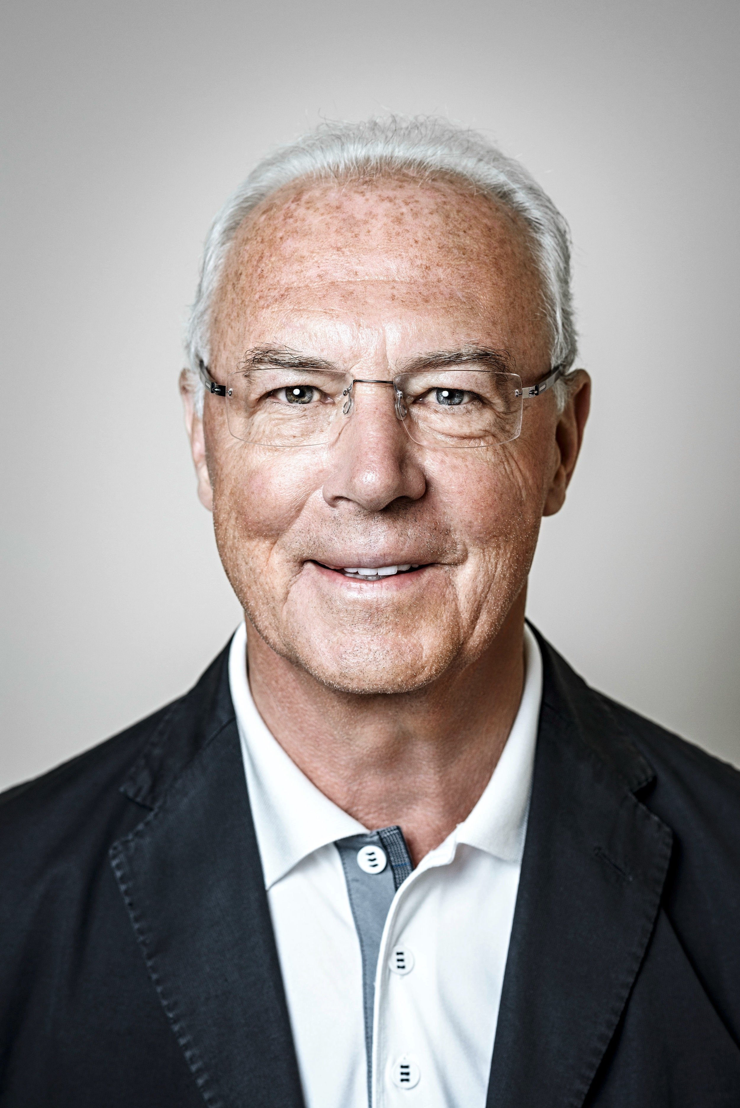 Porträt von Franz Beckenbauer mit randloser Brille und dunklem Sakko vor hellem Grund – Retusche Bildergut.