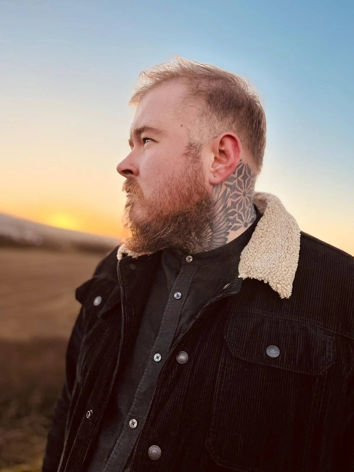 A man with a beard and tattoos on his neck standing outdoors during sunset.