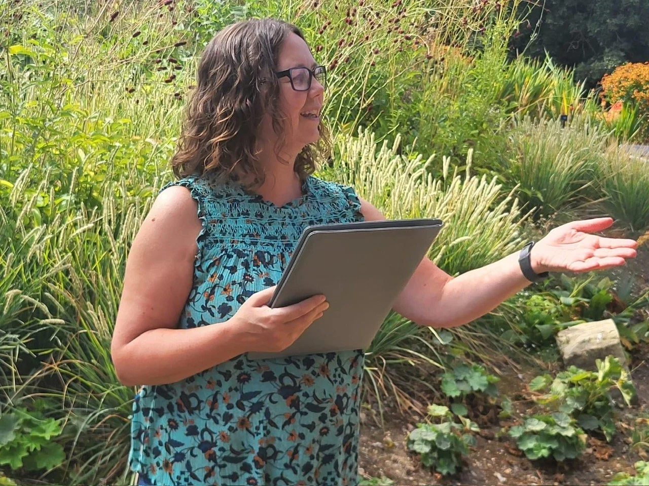 A woman wearing glasses, a blue sleeveless top with a floral pattern, and a wristwatch is standing outdoors among green plants and tall grasses, holding a notebook in one hand and gesturing with her other hand.