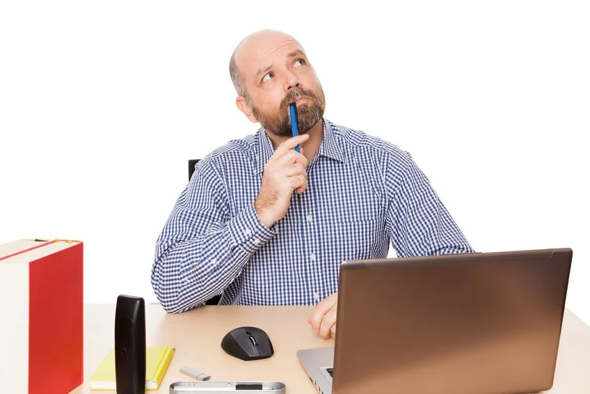 Ein Mann sitzt an einem Schreibtisch mit einem Laptop, einem Smartphone, einer Maus, einem Notizbuch und einem roten Geschenk. Er hat einen Bleistift im Mund und überlegt nachdenklich. Modul 1 Theorie bei Tinnitus.