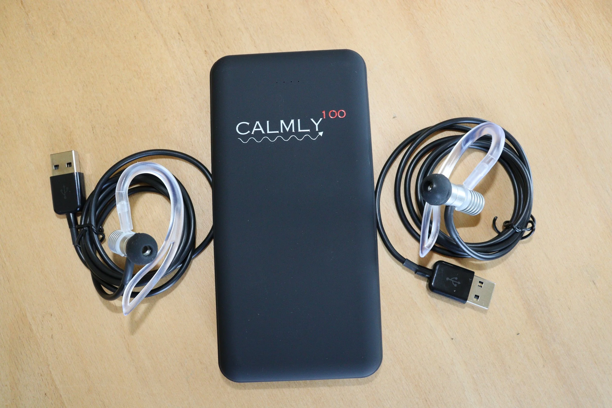 Ein schwarzes Gerät mit der Aufschrift 'CALMLY 100' liegt auf einem Holztisch. Es ist von mehreren Kabeln umgeben, inklusive einem USB-Kabel und einem silbernen Ohrhörer.