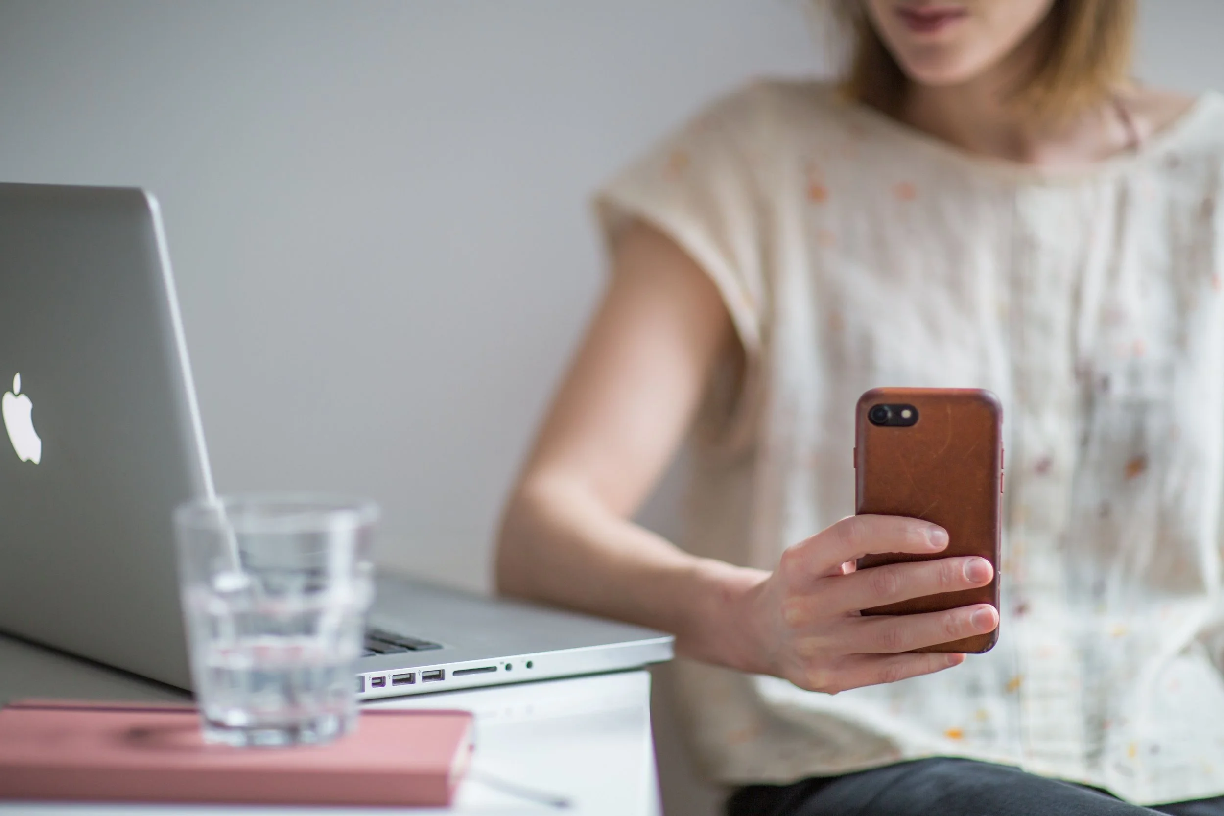 Frau hält ein Smartphone in der Hand, während sie an einem Laptop sitzt, im Hintergrund eine weiße Wand. Melde Dich gerne, wenn Du Fragen hast zum Tinnitus Online Kurs und zum Heimlaser.