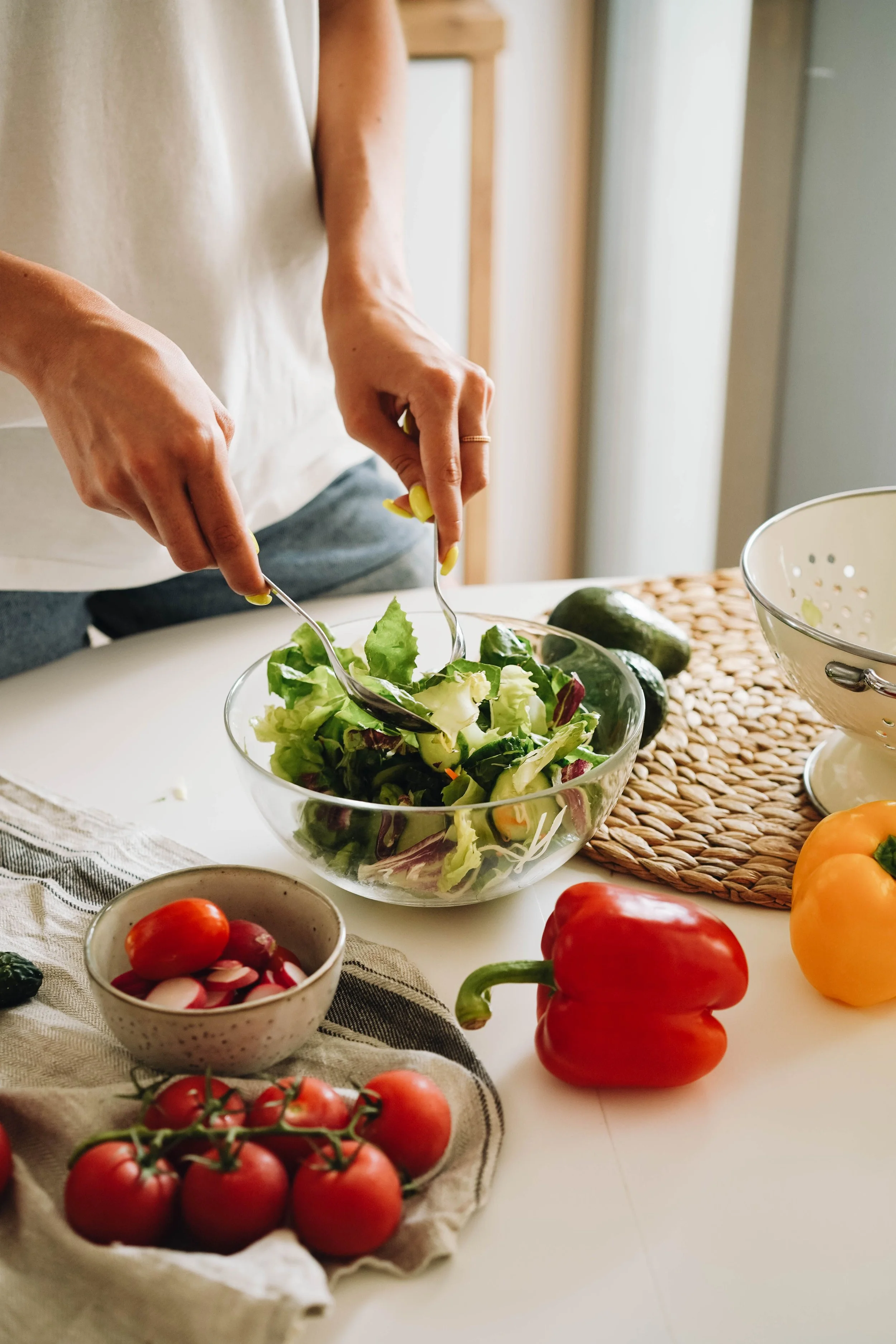 Person bereitet einen frischen Salat mit grünem Salat in einer Glasschüssel. Verschiedene Gemüsesorten, darunter Tomaten, Paprika, Gurken und Radieschen, sind um den Tisch verteilt. Modul 9. Ernährung bei Tinnitus.