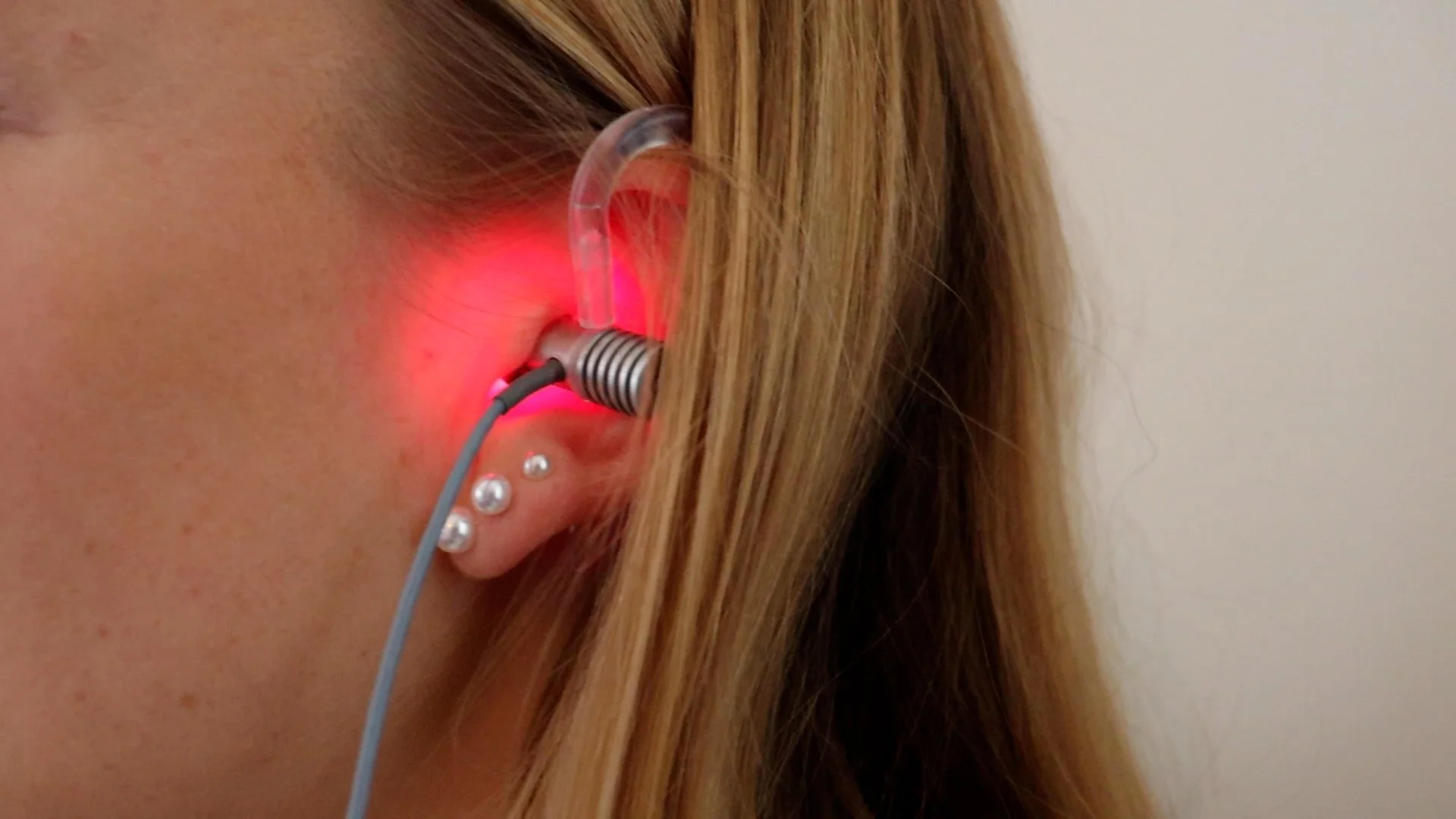 A close-up of a person's ear with a hearing aid emitting a red light, accompanied by three piercings with metallic balls.