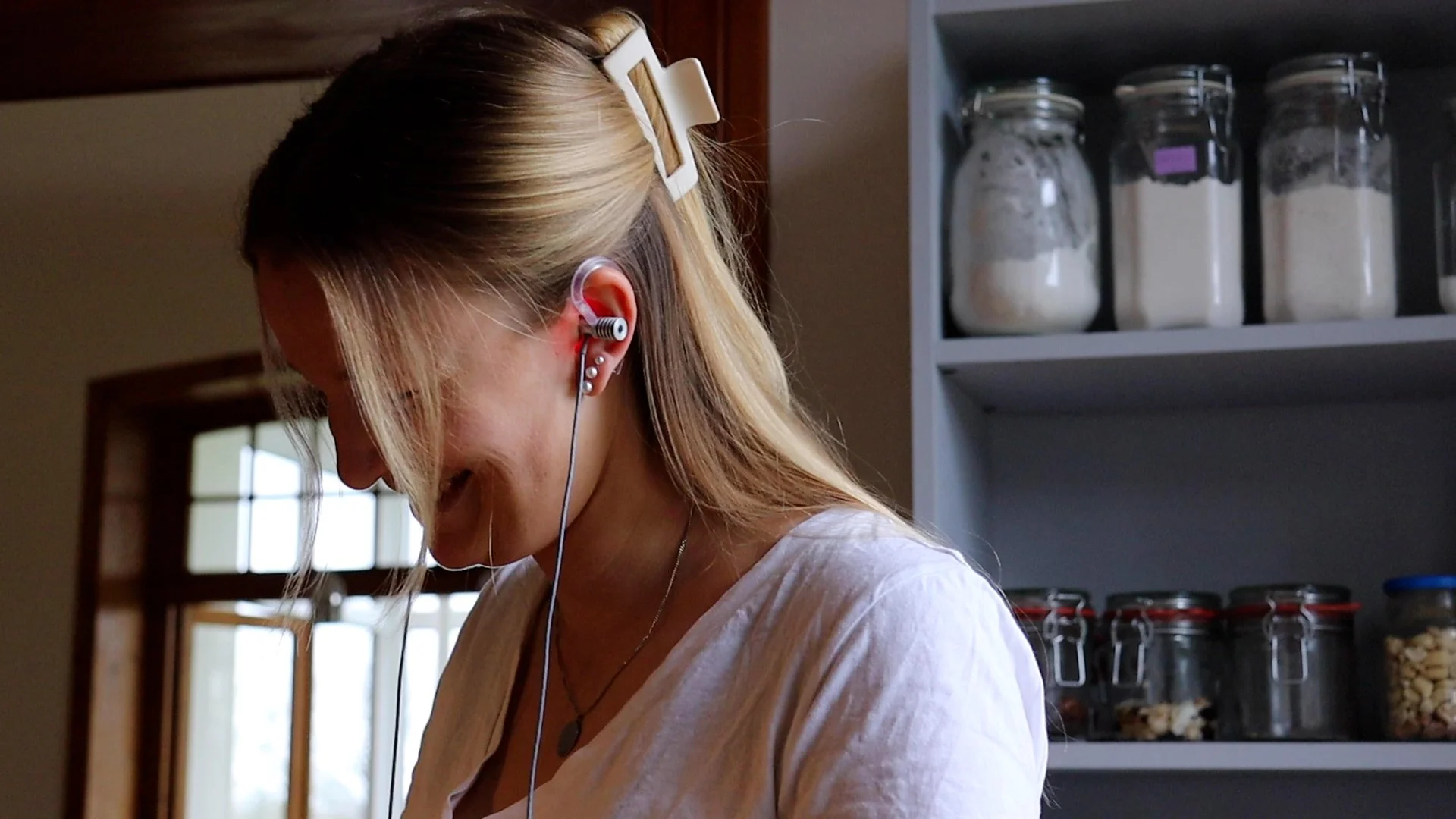 Eine lächelnde Frau mit langem blondem Haar trägt einen Hörgerät und hört Musik, steht in einer Küche vor einem Regal mit Gläsern, die mit Mehl, Zucker und Keksen gefüllt sind.