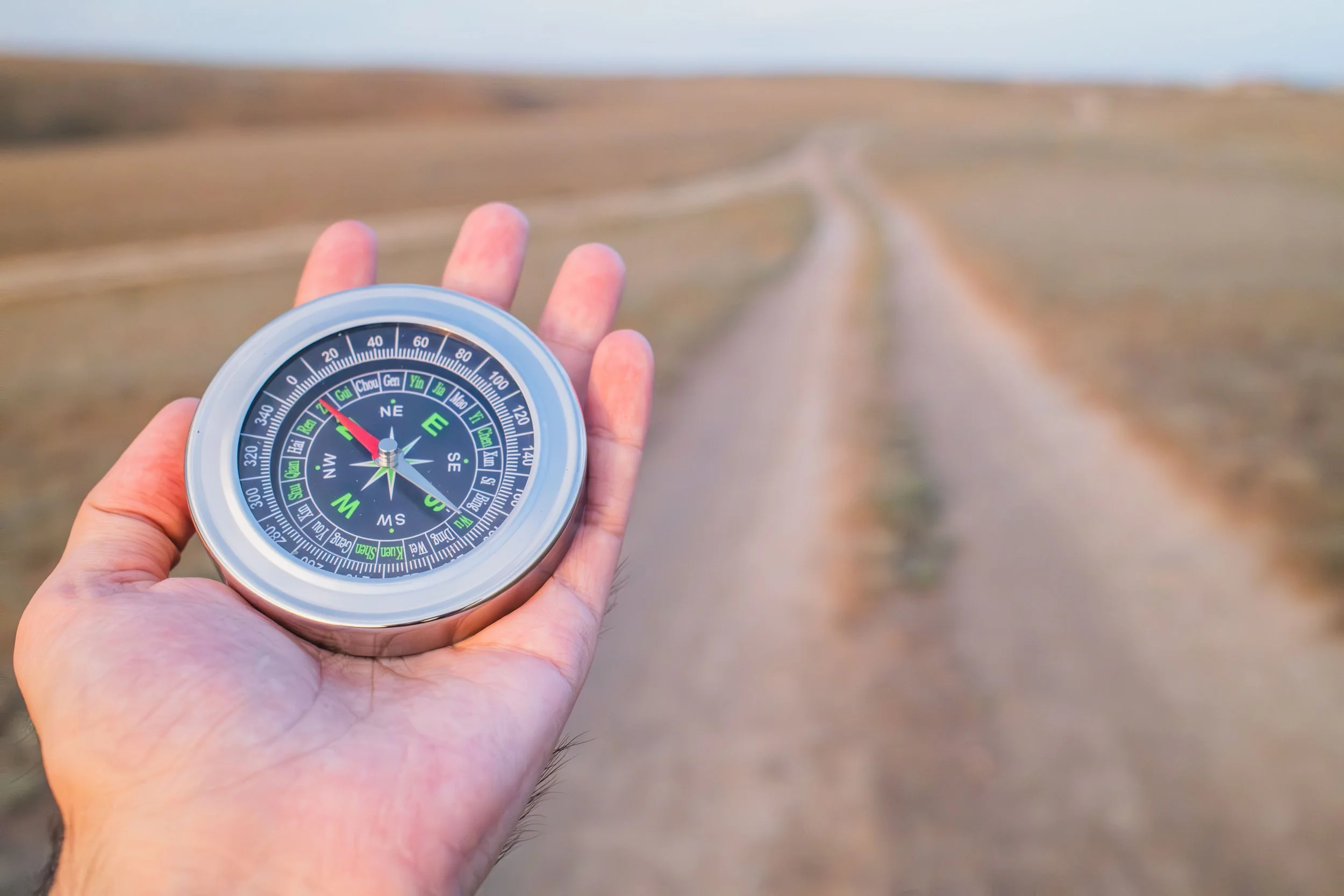 Nahaufnahme einer Hand, die einen Kompass hält, mit einer unbefestigten Straße, die in die weite offene Landschaft führt, im Hintergrund. Modul 13. Abschluss und Orientierung bei Tinnitus.