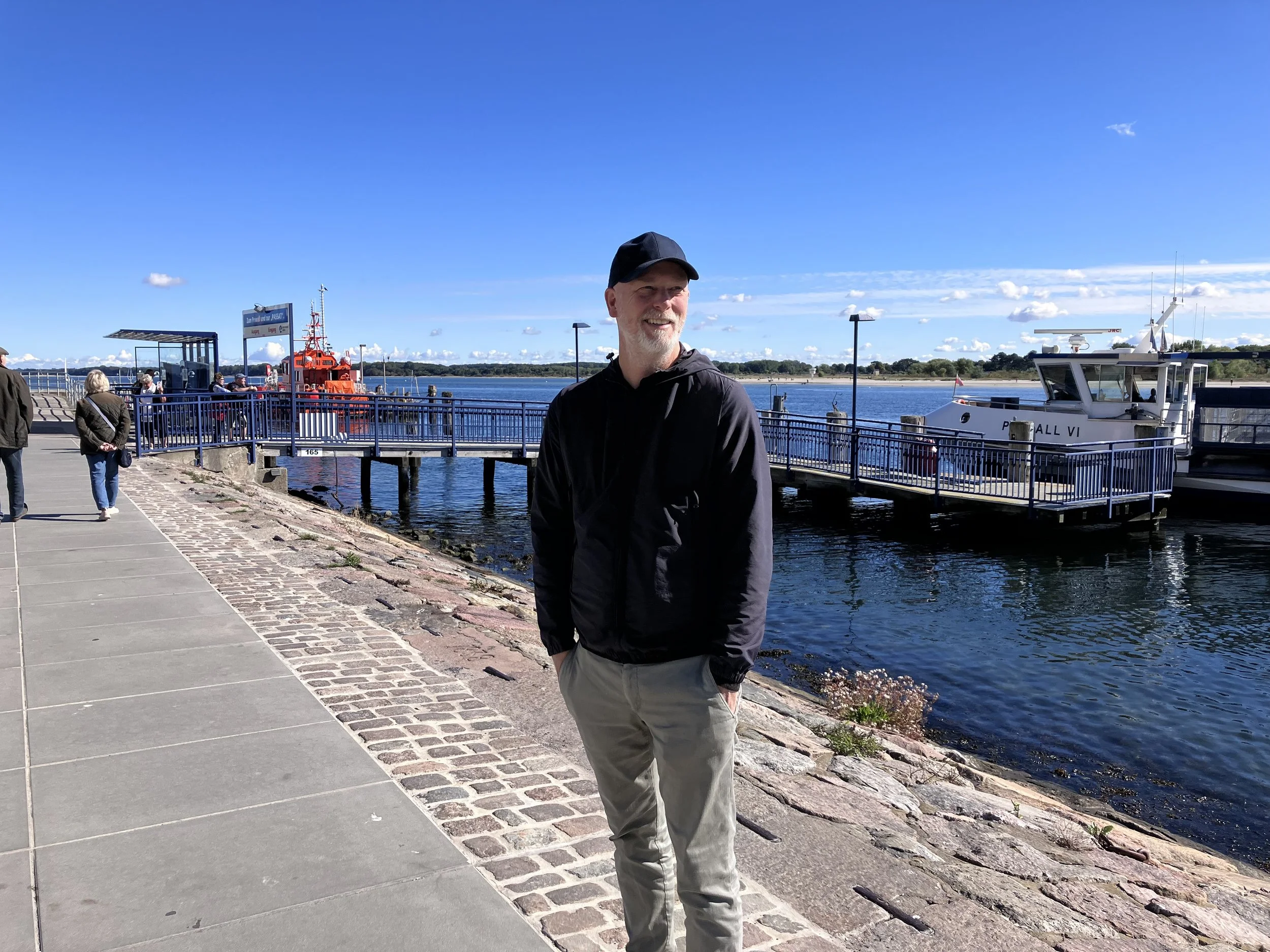 Ein Mann mit grauem Bart, schwarzer Mütze und schwarzer Jacke steht an einem Hafen bei Sonnenschein, im Hintergrund Boote und Wasser, einige Leute laufen entlang der Uferpromenade. Mehr über HEALTH Services Robert Kroll.