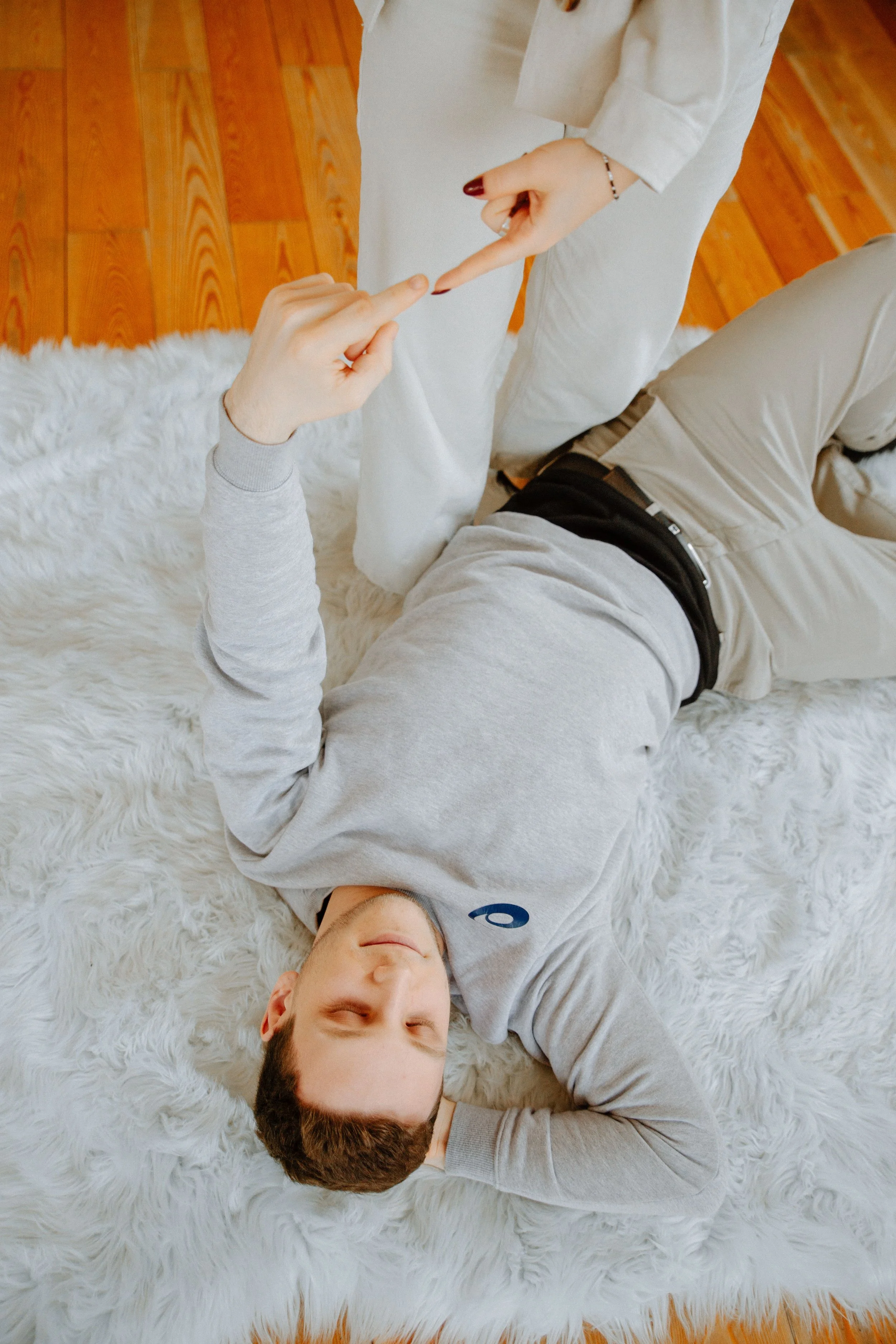 Ein junger Mann liegt entspannt auf einem weißen Fellteppich, trägt eine graue Kapuzenjacke und beige Hosen, und scheint zu lächeln. Der Raum hat Holzboden. Modul 6 Intensive Körperübungen bei Tinnitus.