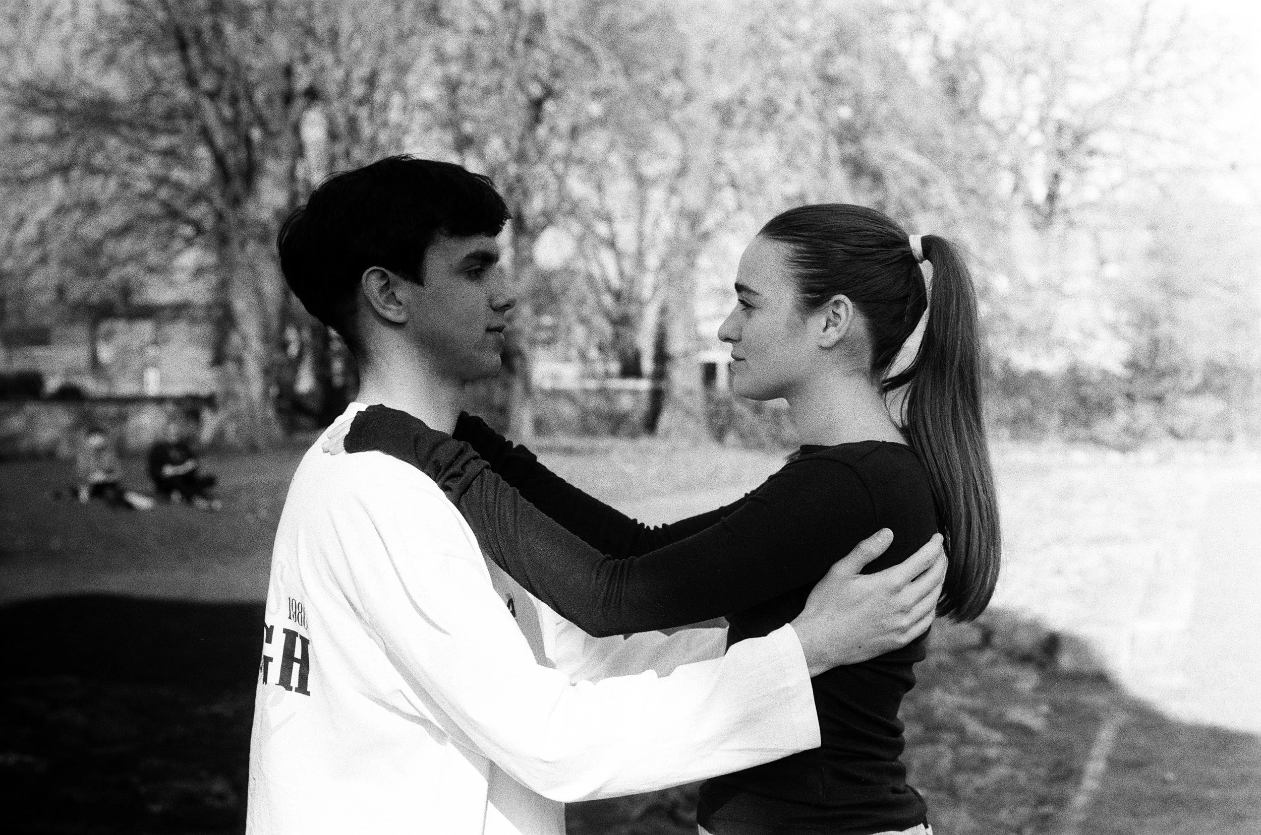A black-and-white photo of a young man and woman standing closely face to face outdoors, holding each other's shoulders. The woman has a ponytail and is wearing a long-sleeve shirt, while the man has short hair and is wearing a T-shirt. They are looking into each other's eyes, with blurred trees and people in the background.