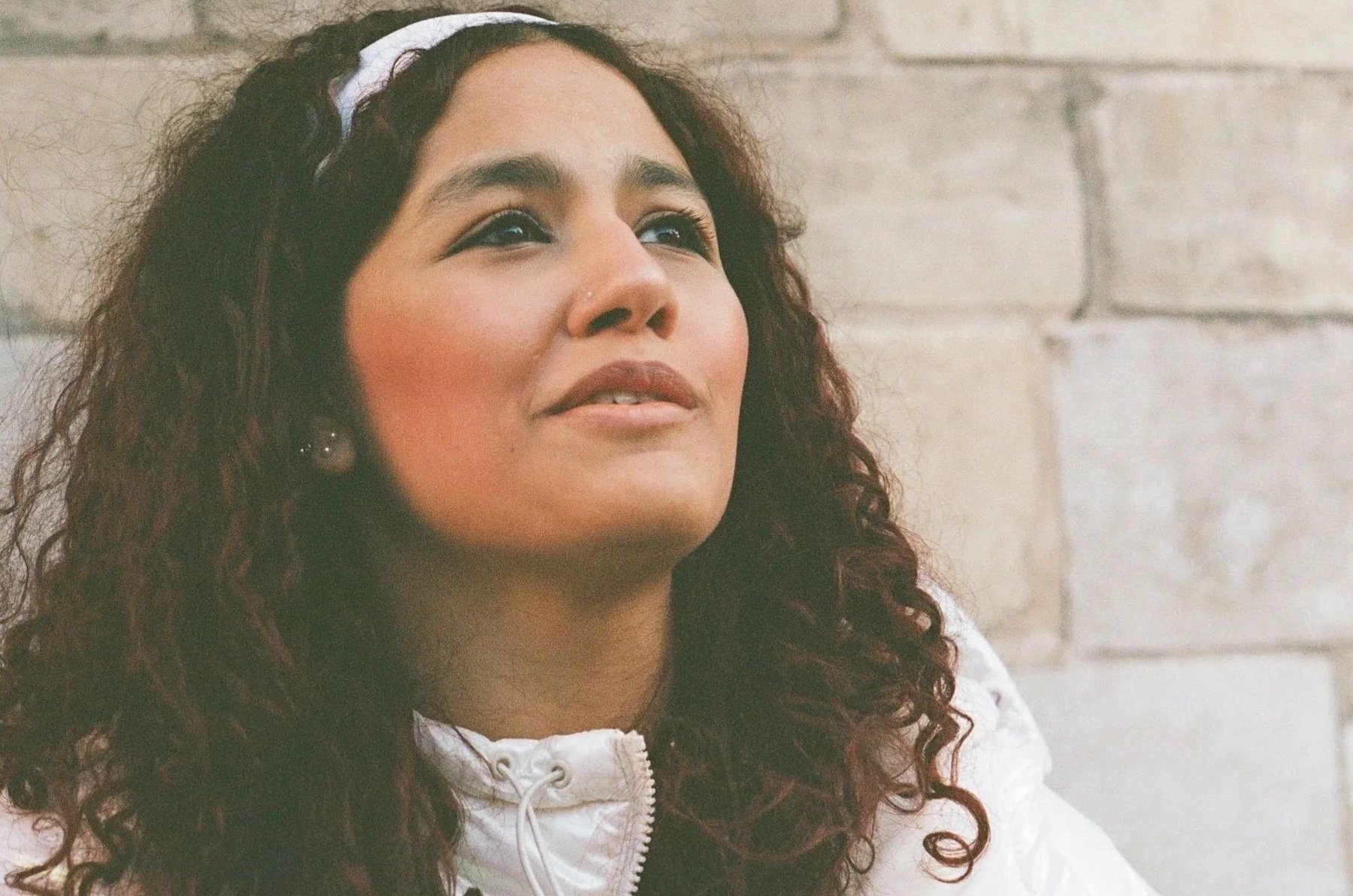 Close-up of a woman with curly hair wearing a white jacket, looking upwards with a slight smile, standing in front of a stone wall.