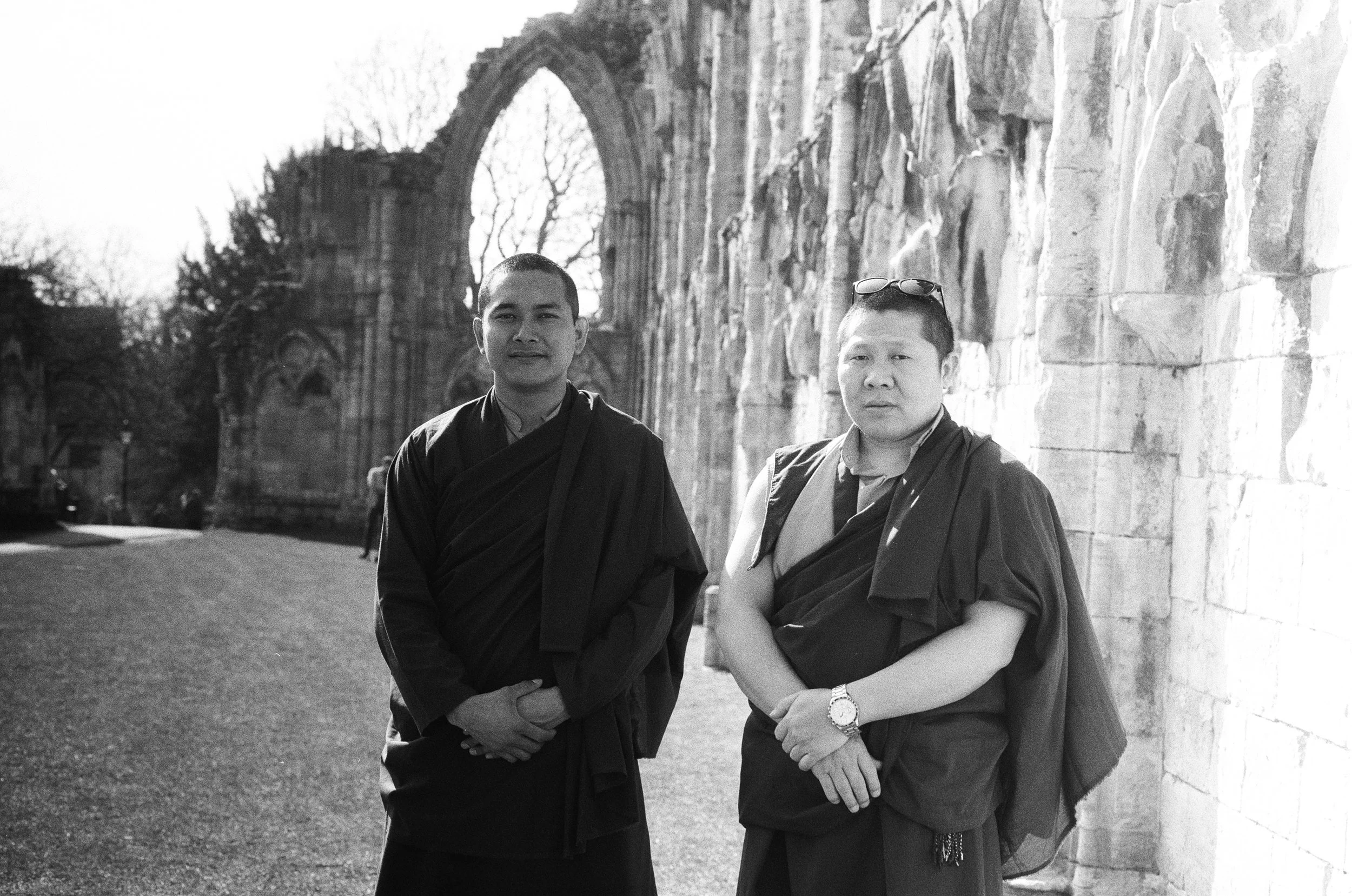 Two monks in robes standing outdoors near an ancient stone structure with arches, with trees in the background.