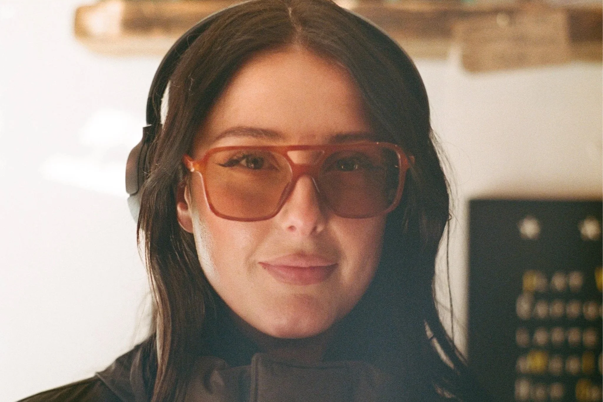 Close-up of a woman with long brown hair, wearing large sunglasses and headphones, smiling slightly, with a blurred bookshelf in the background.
