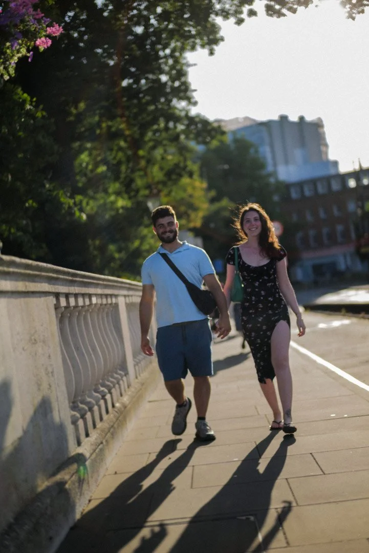 A young couple walking and holding hands on a city sidewalk during late afternoon or early evening, with buildings and trees in the background.