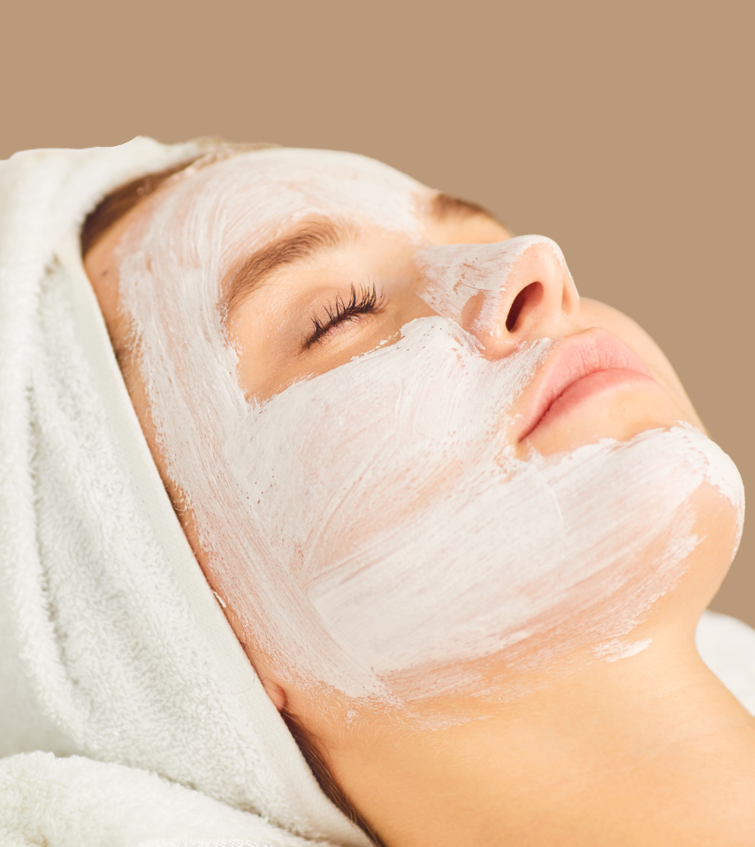 Mujer recibiendo un tratamiento facial con una máscara blanca, con los ojos cerrados y envuelta en una toalla blanca, en un ambiente de spa.
